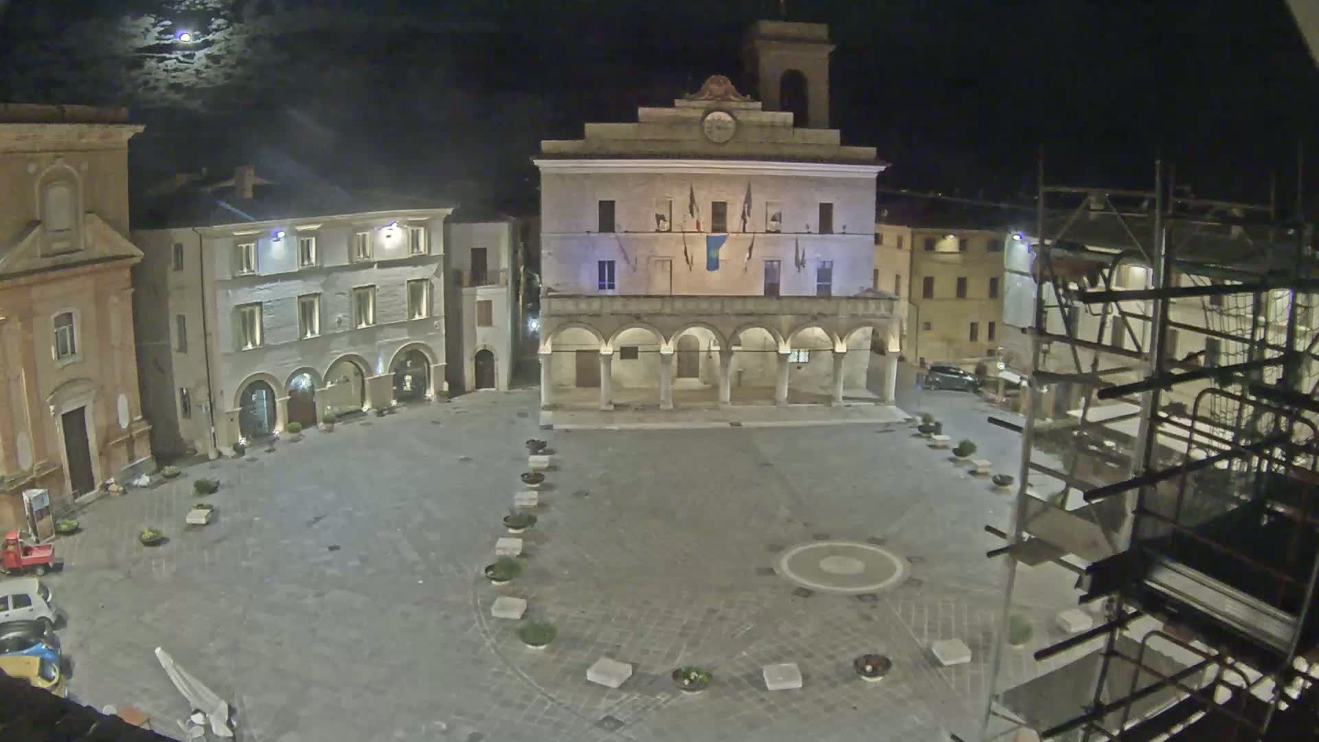 A moonlit town square at night features a large, arcaded building, surrounded by smaller buildings, with scaffolding visible on the right.