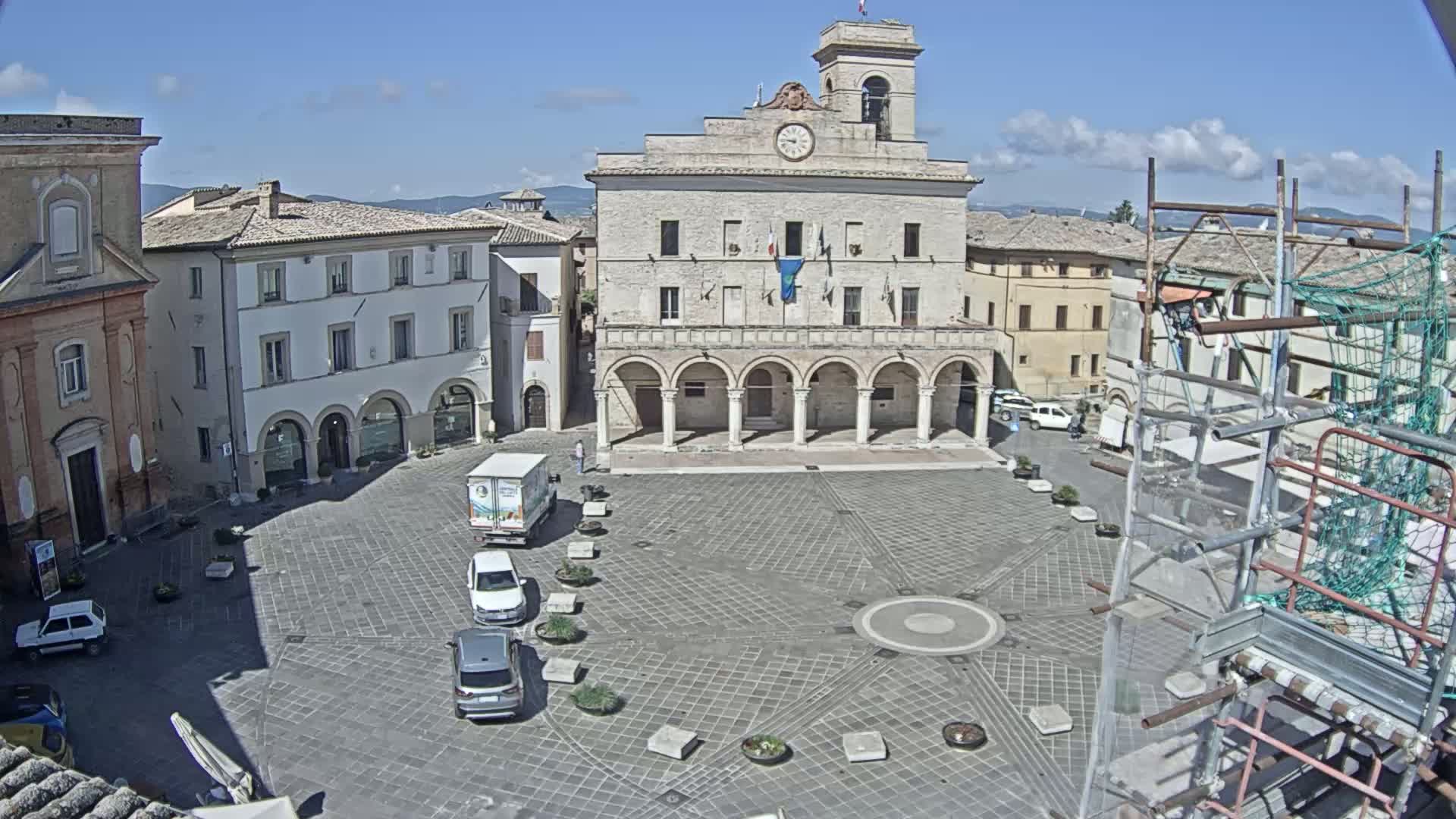 A town square with a large stone building, several smaller buildings, a few cars, and scaffolding under a mostly sunny sky.