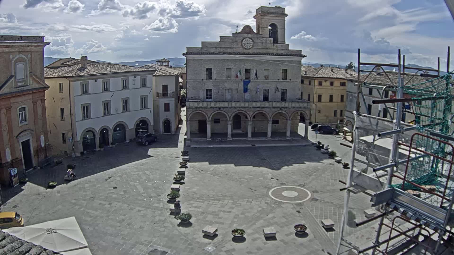 A large town square under partly cloudy skies is centered on a large, arched stone building, with several other buildings surrounding the square and scaffolding visible in the bottom right corner.