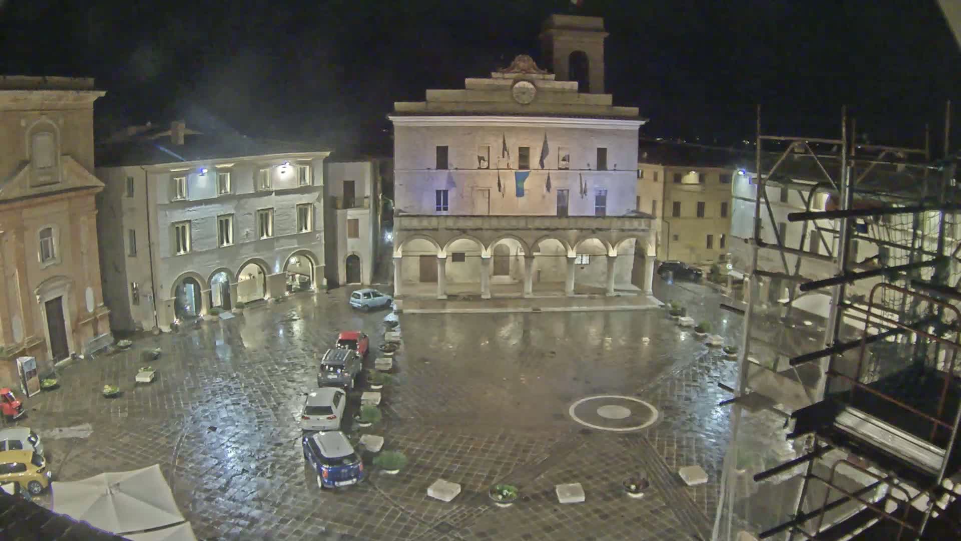 A rain-slicked town square at night, centered around a large building with arched entryways, surrounded by other buildings and a few parked cars.
