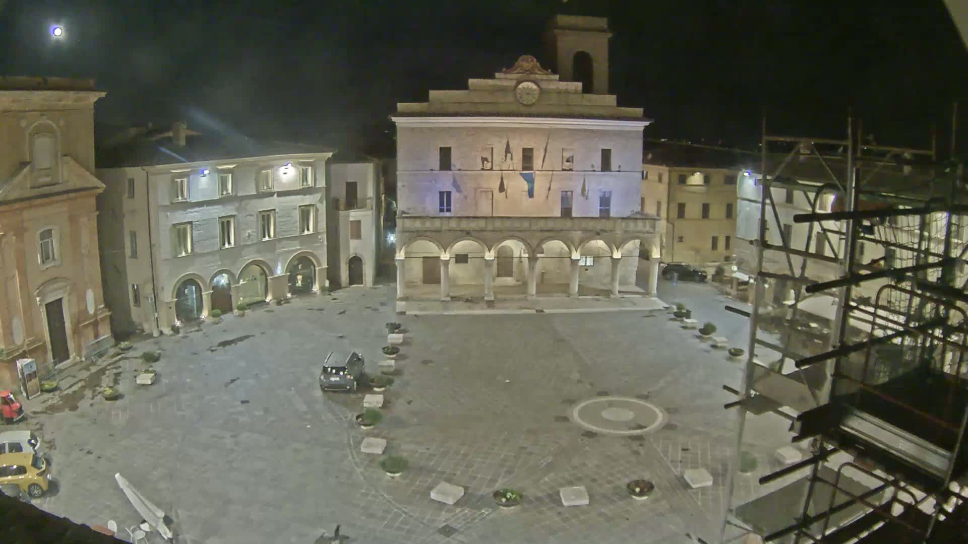 At night, under a partly cloudy sky, a town square is shown with a large building featuring arches, several smaller buildings, and scaffolding to the right.