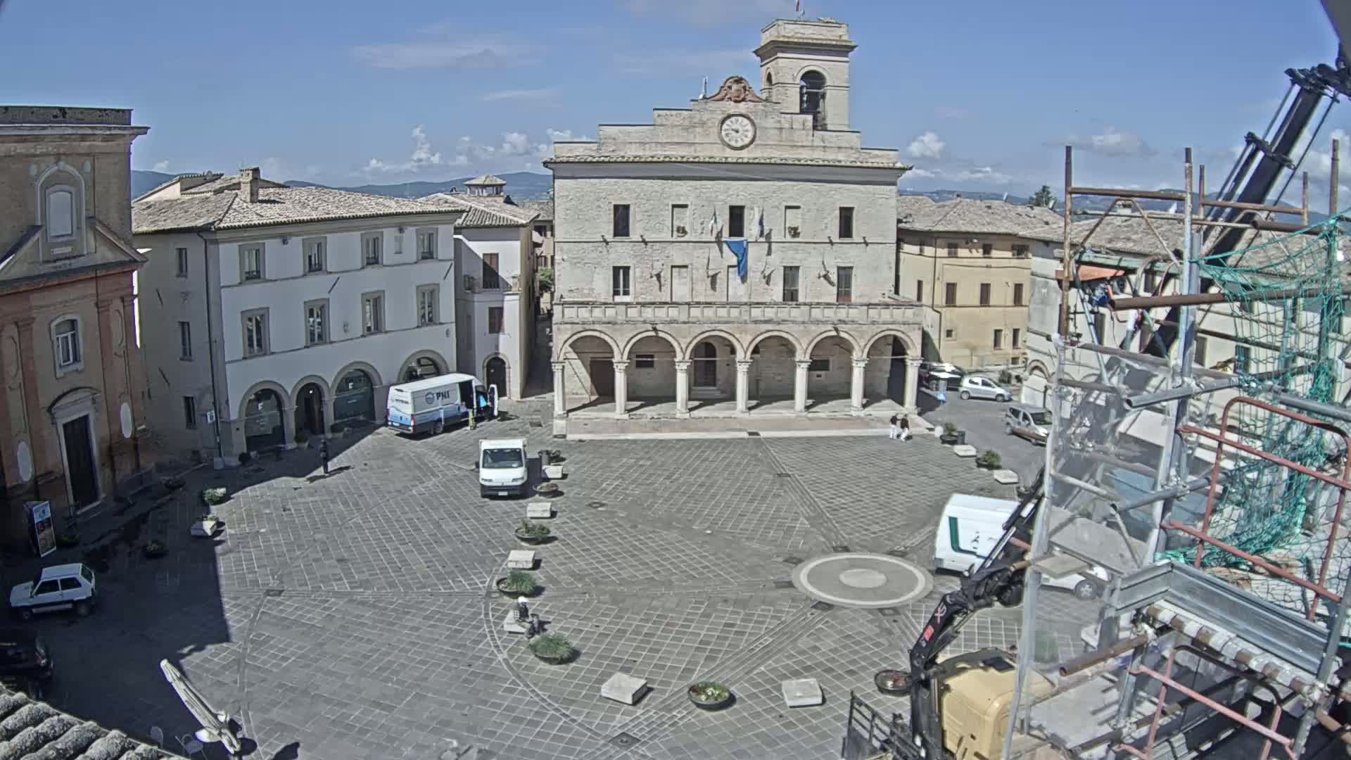 A sunny day reveals a town square with a large building featuring arched entryways, surrounded by other buildings, vehicles, and scaffolding.
