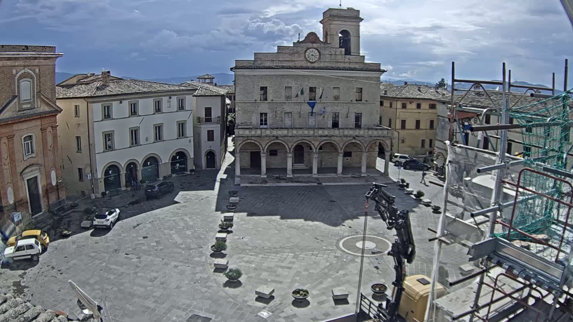 A mostly empty town square with a large building featuring arched entryways and a clock tower, under a partly cloudy sky.
