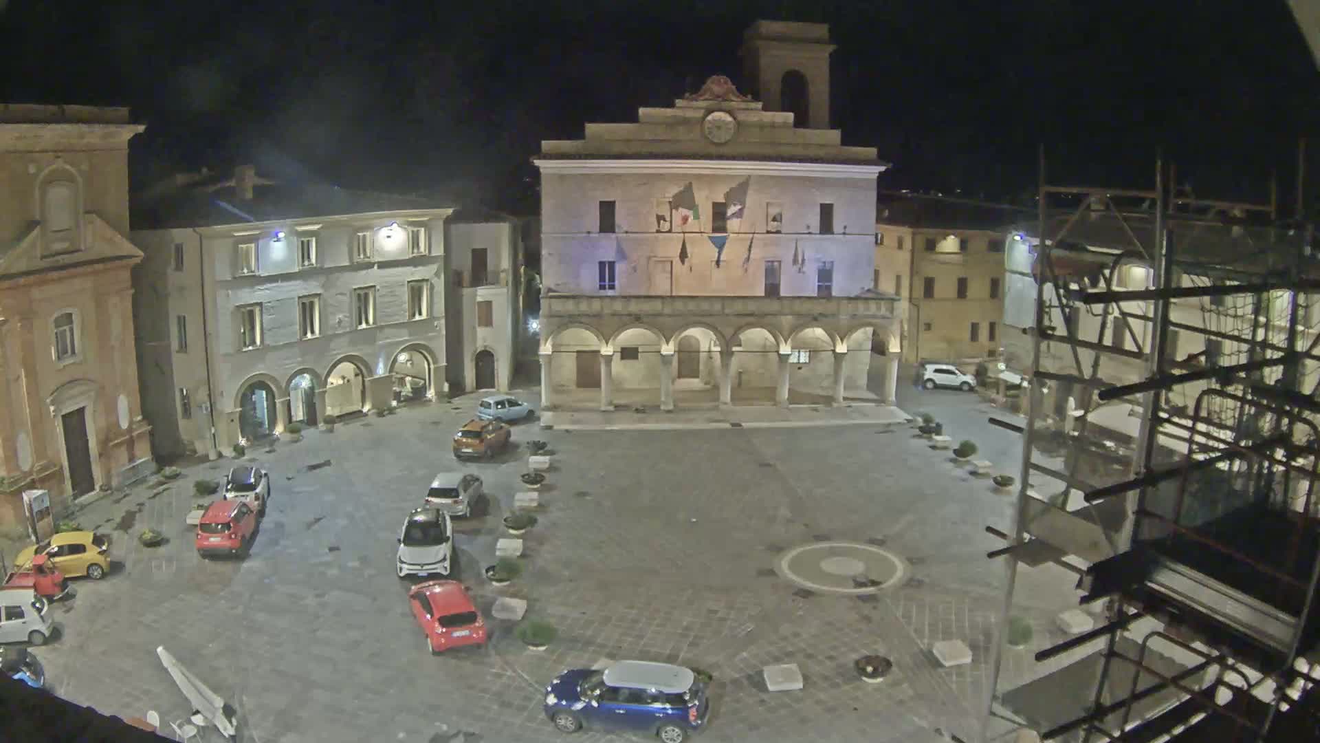 A nighttime view of a town square with several cars parked, a large building with arches, and scaffolding on a building to the right.
