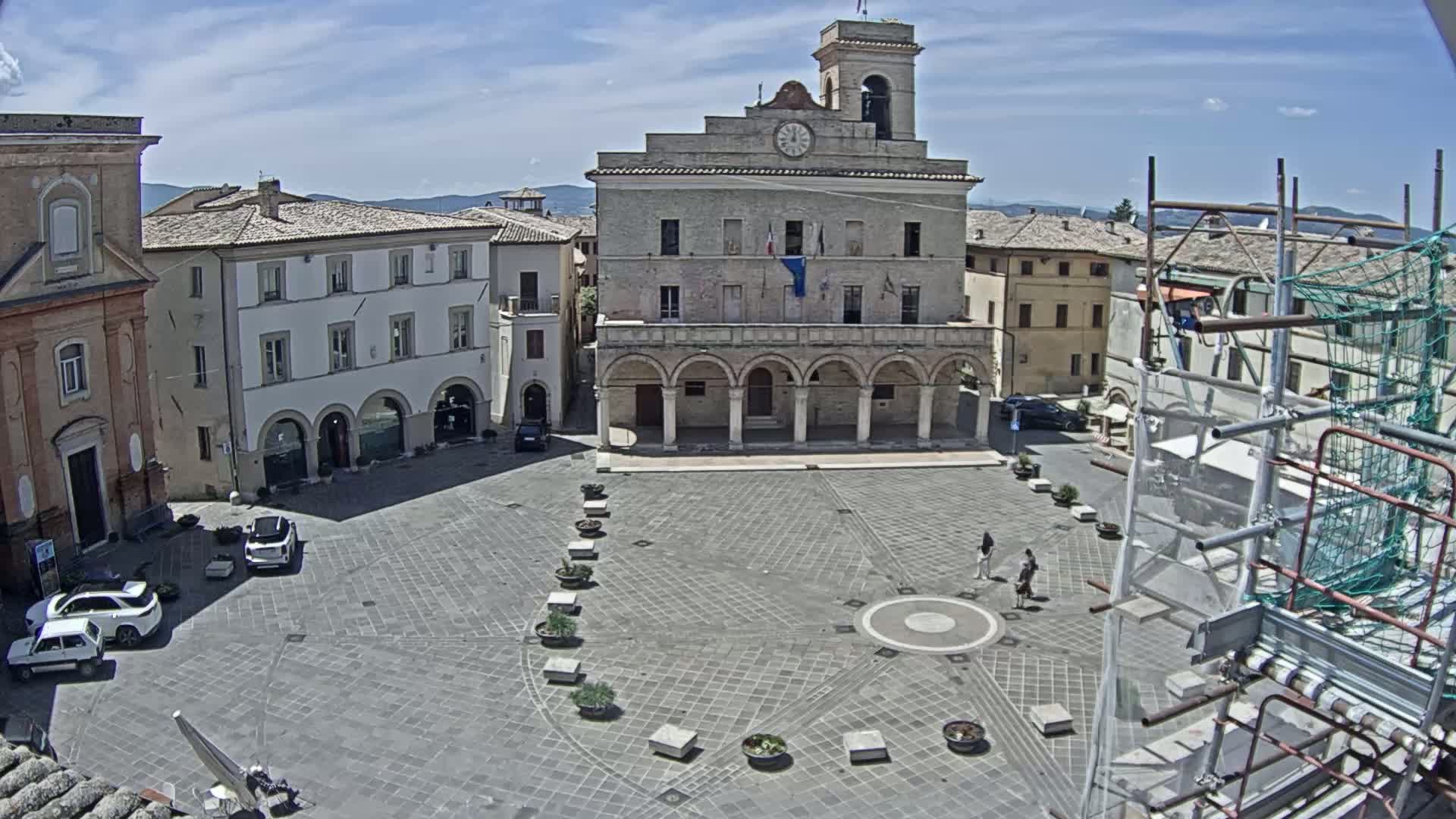 A large town square with a prominent stone building, surrounded by other buildings, under a sunny sky.