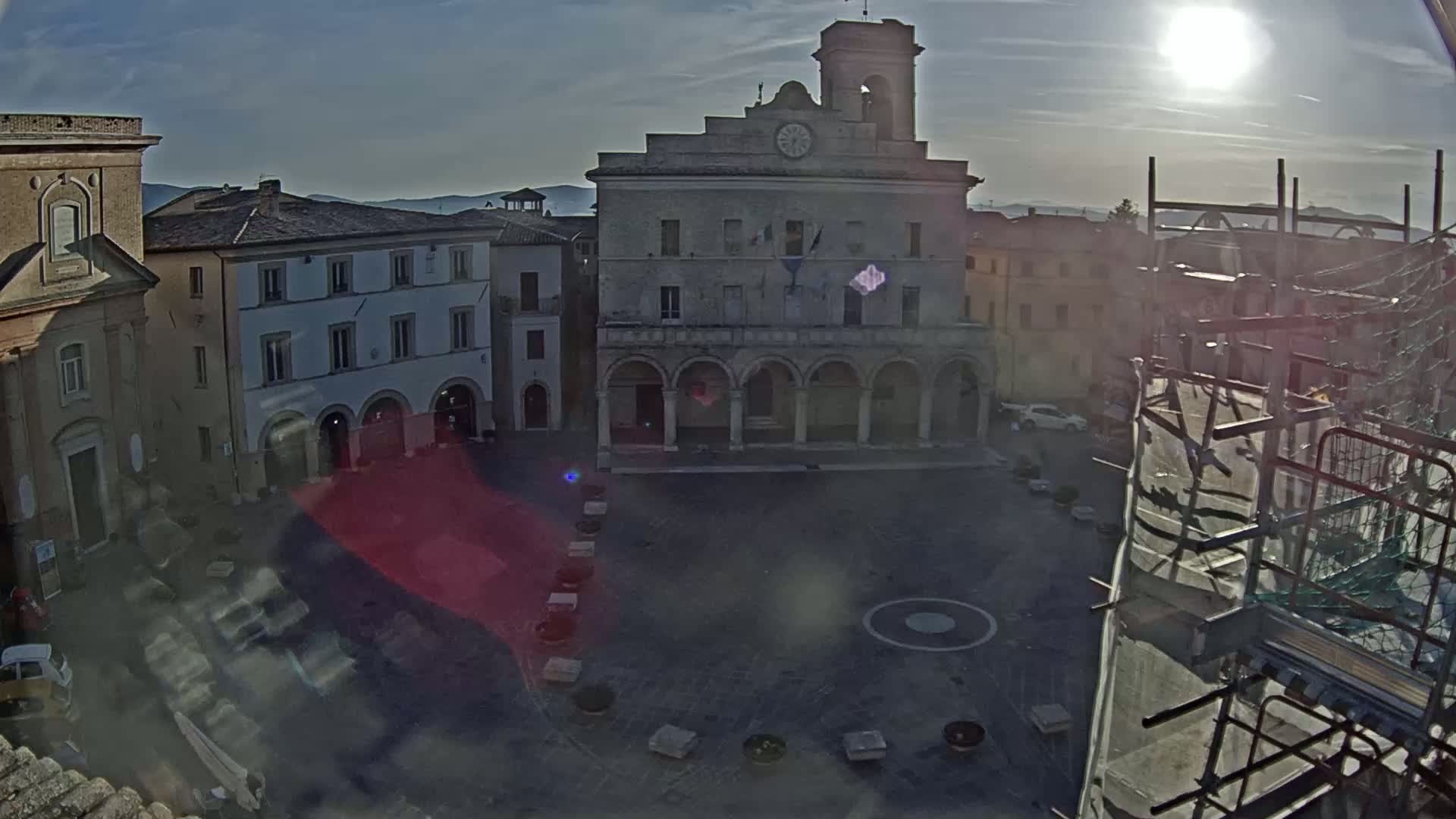 A sunlit town square, featuring a large stone building with arched entryways, is surrounded by other buildings under a mostly clear sky.