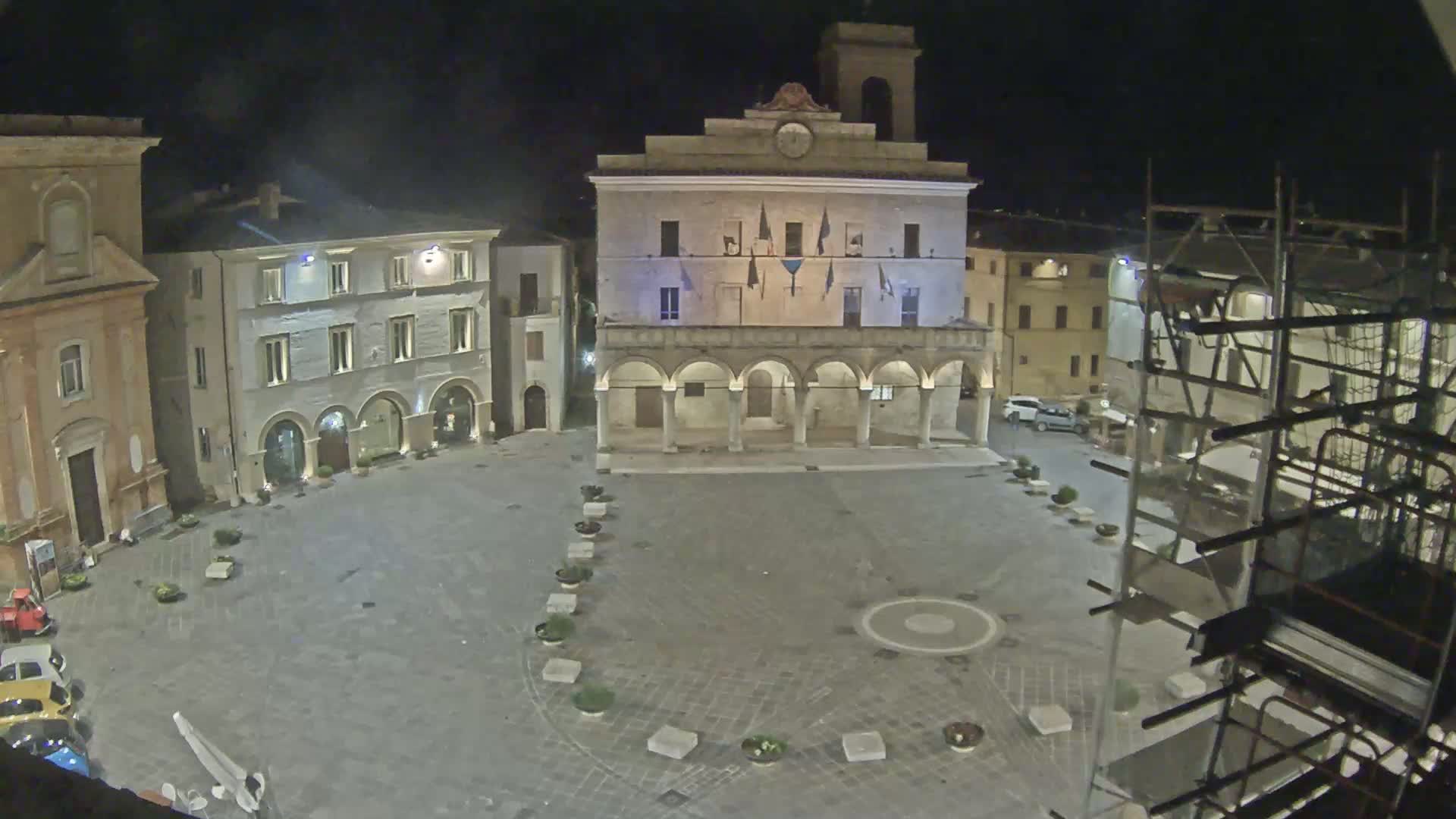 At night, a town square is seen, with a large building featuring arched entryways at its center, surrounded by other buildings and scaffolding.
