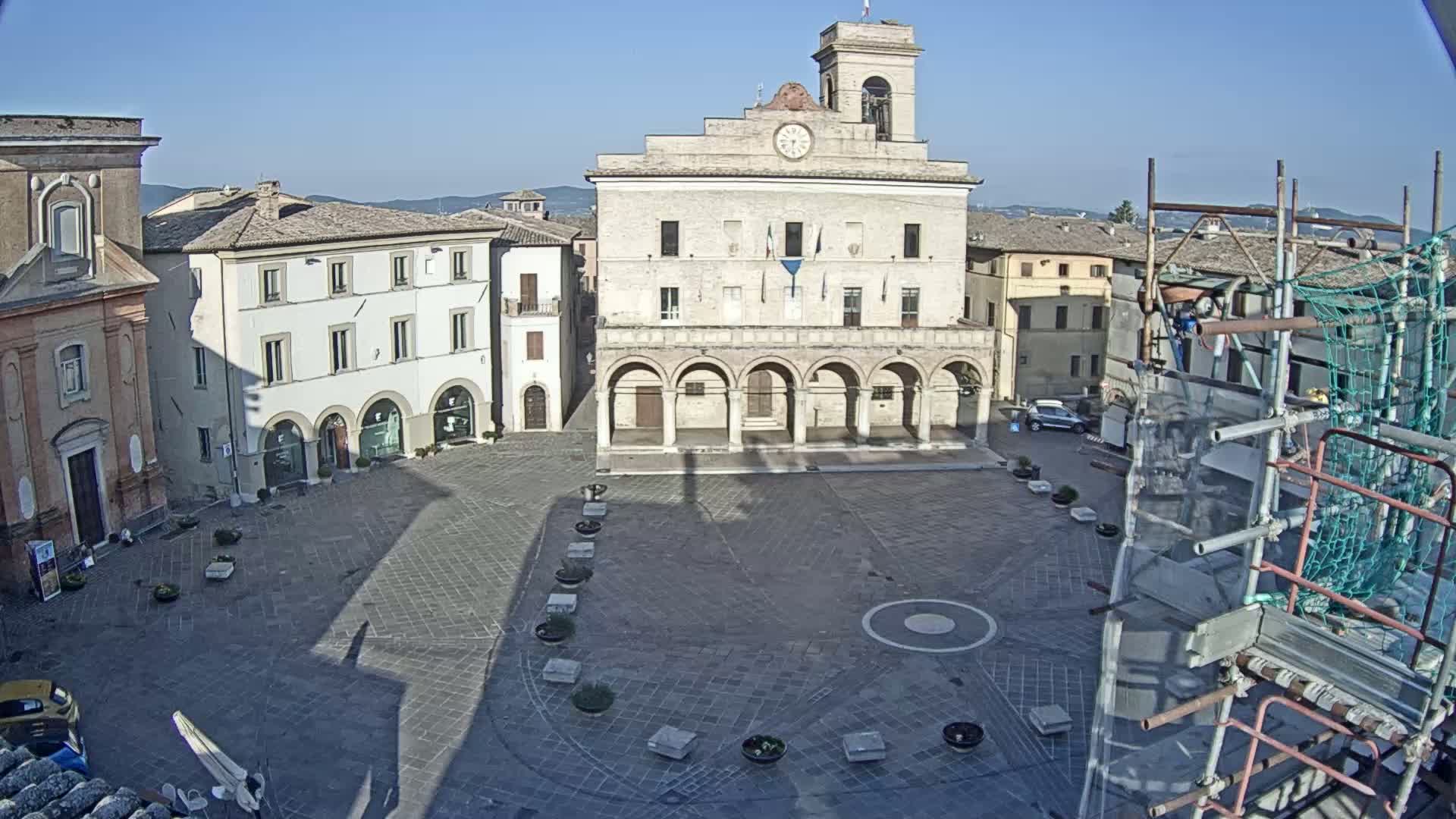At night, a town square is seen, with a large building featuring arched entryways at its center, surrounded by other buildings and scaffolding.