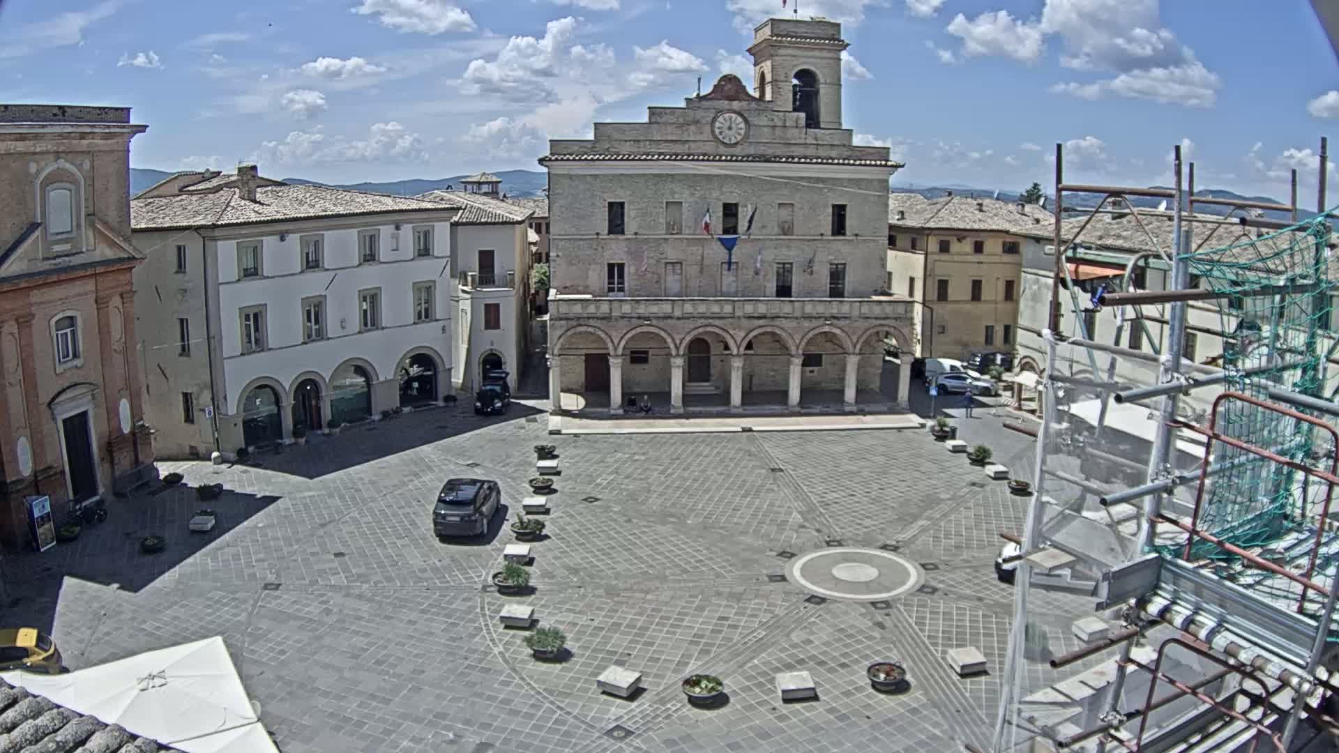 A town square with a large stone building, several other buildings, and a few cars, under a mostly sunny sky.