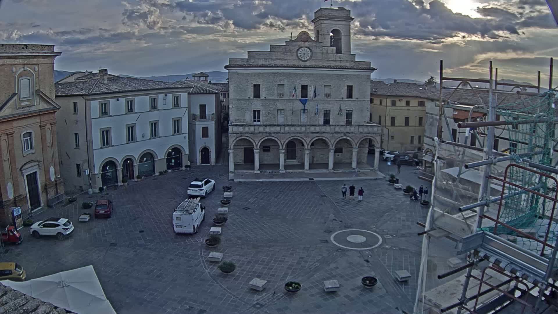 A town square with a large stone building, several smaller buildings, and a few cars under a partly cloudy sky.
