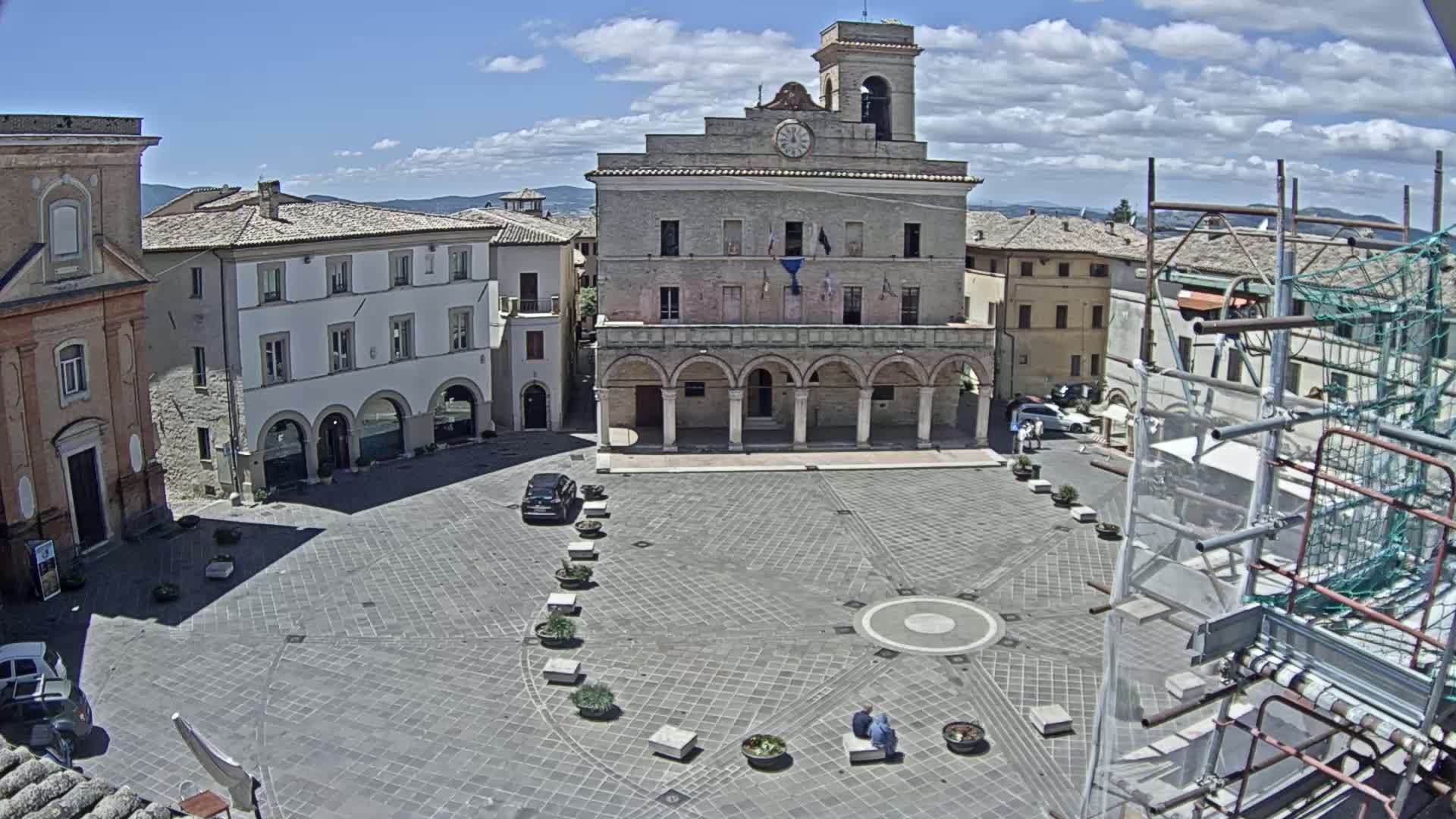 A town square, under a mostly sunny sky with some clouds, features a large stone building with arched entryways, surrounded by other buildings, and contains a few cars and people.