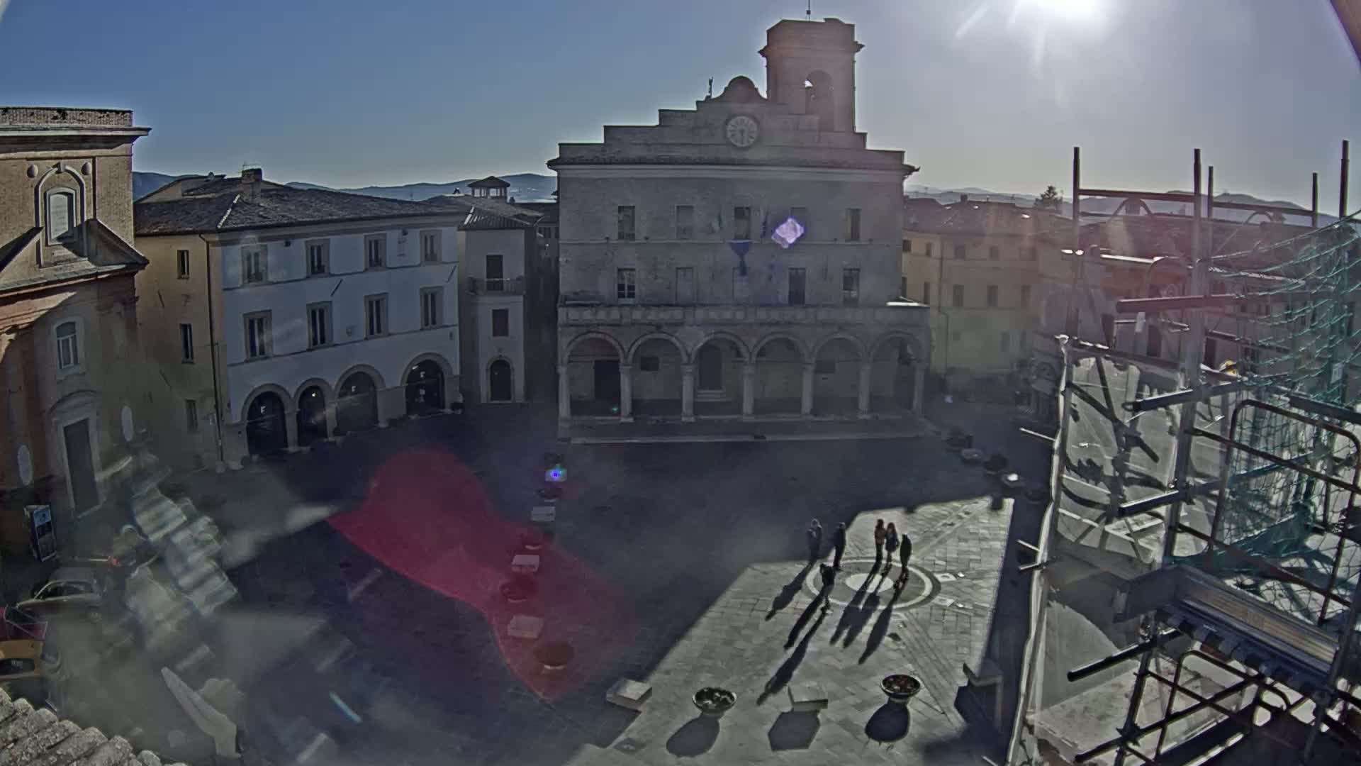 A sunny day reveals a town square with a large stone building, several smaller buildings, and a few people casting shadows on the paved ground.