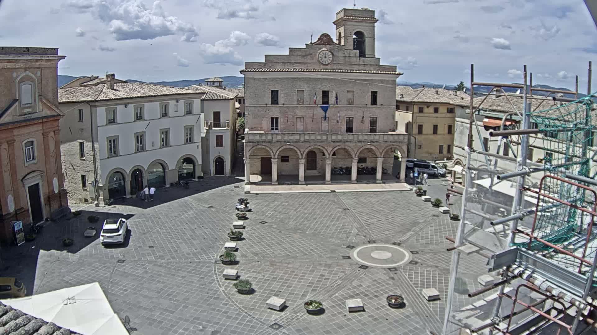 A large, paved town square is centered around a town hall, with several surrounding buildings, under a partly cloudy sky.
