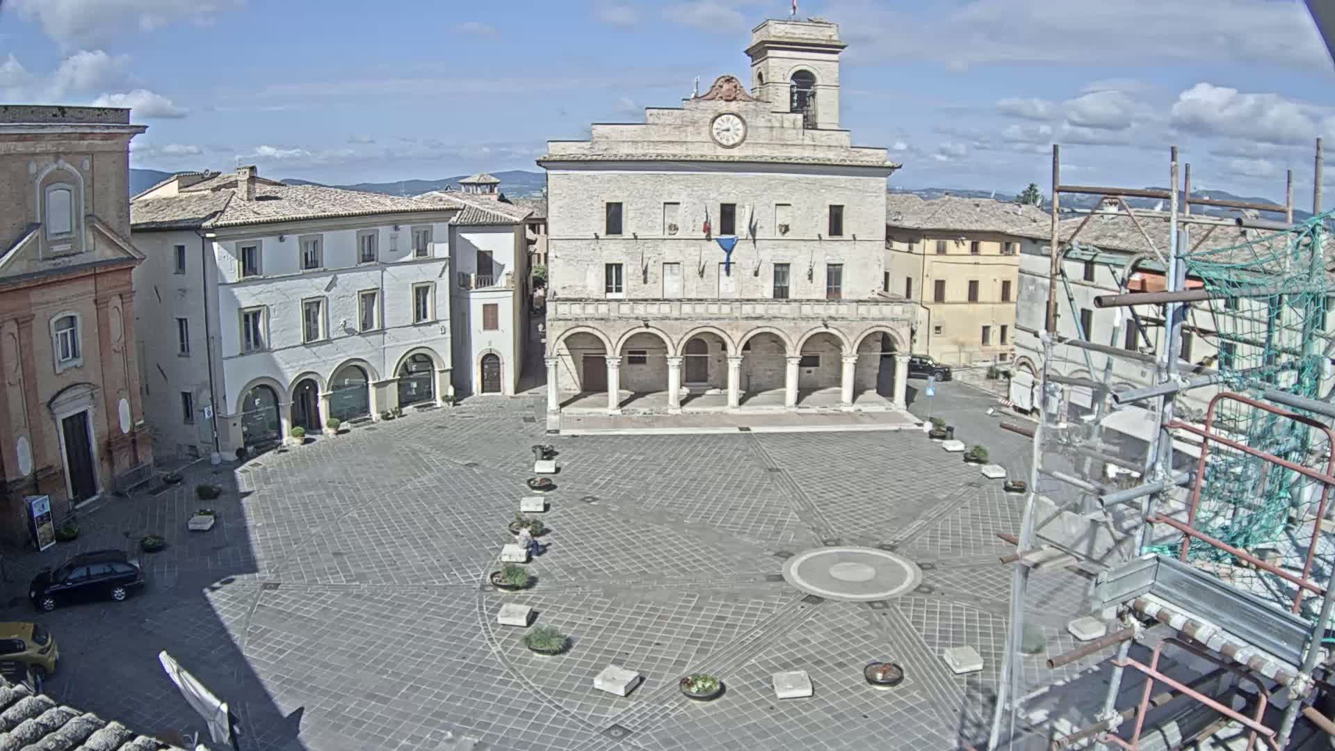 A mostly sunny day reveals a large, paved town square with a grand building featuring arched columns in the center, surrounded by other buildings, and scaffolding on one side.