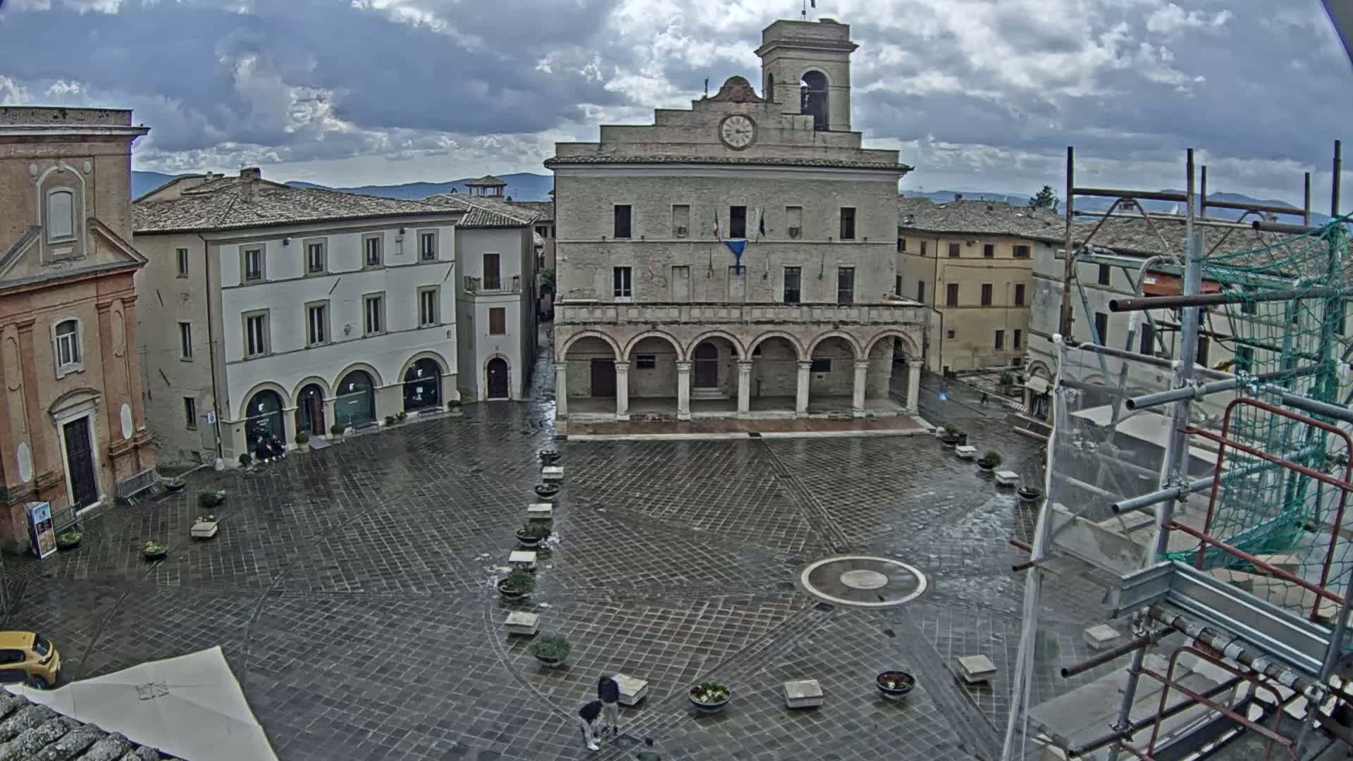 A wet, stone town square is surrounded by buildings, including a large building with arched entries and a clock tower, under a cloudy sky.