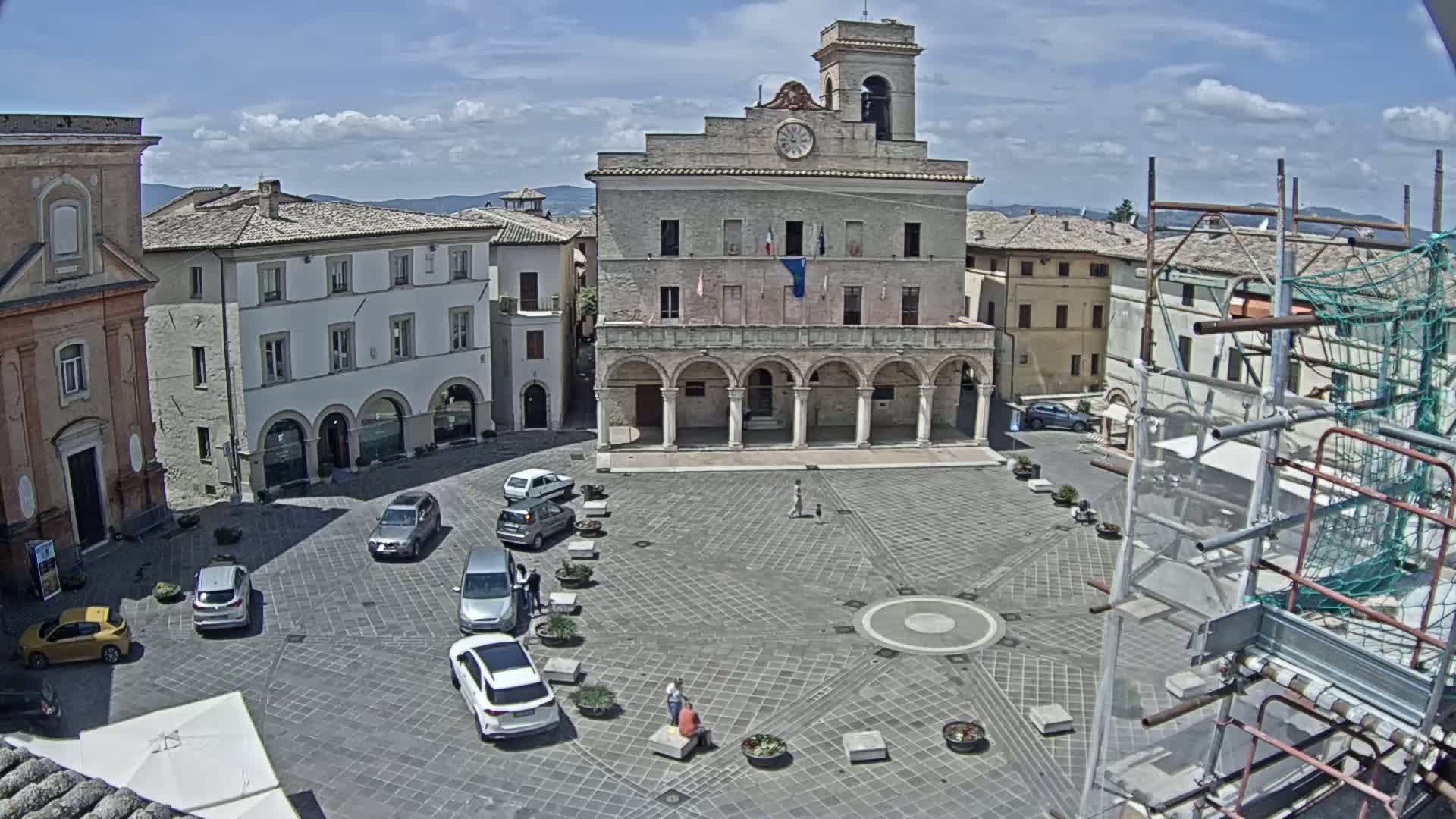 A large town square on a sunny day is surrounded by buildings, including a prominent stone building with arched entryways, and contains several parked cars and a few pedestrians.