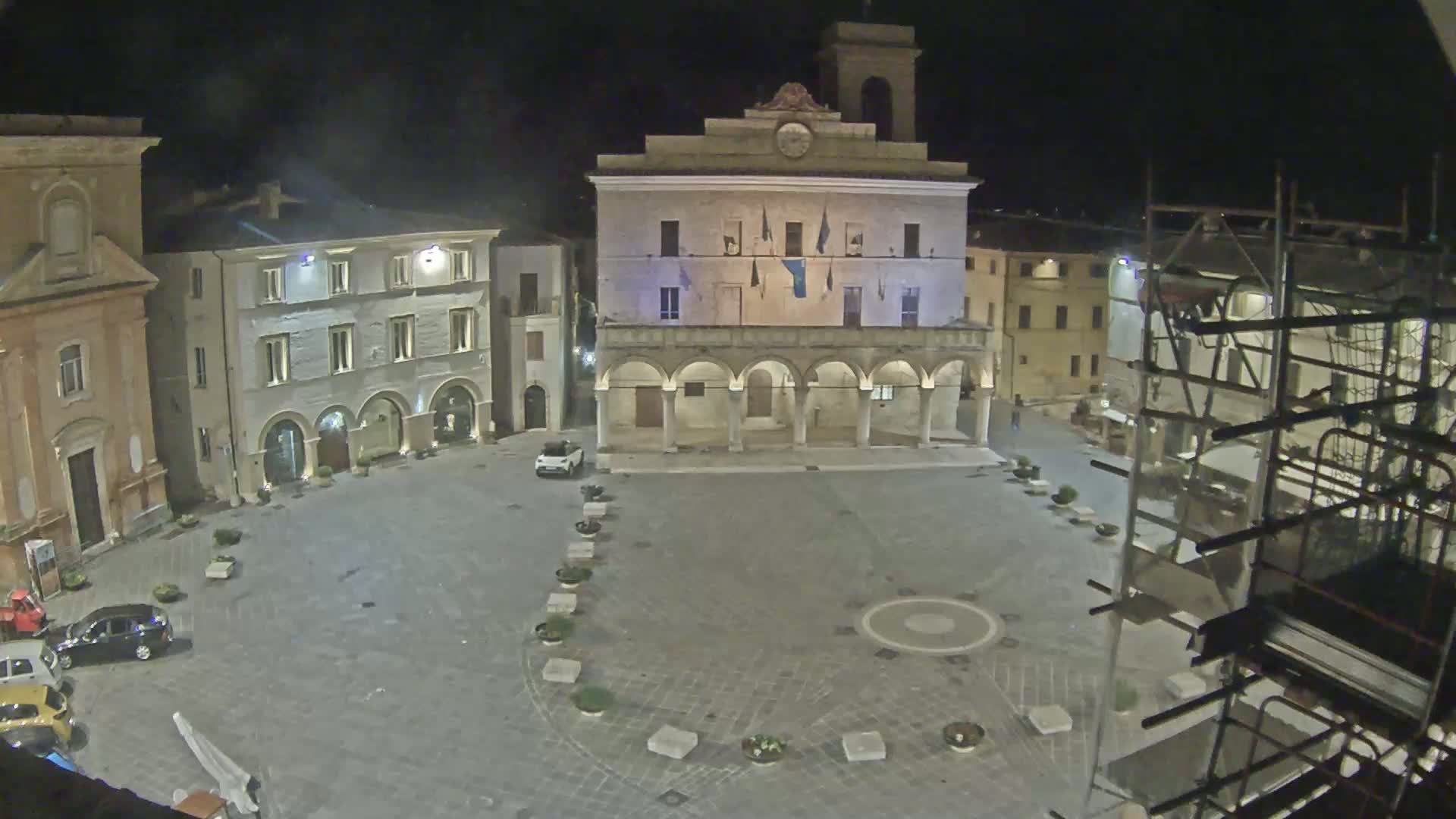 A nighttime view of a town square with several buildings, including a large building with arched entryways, and some cars parked, under a clear sky.
