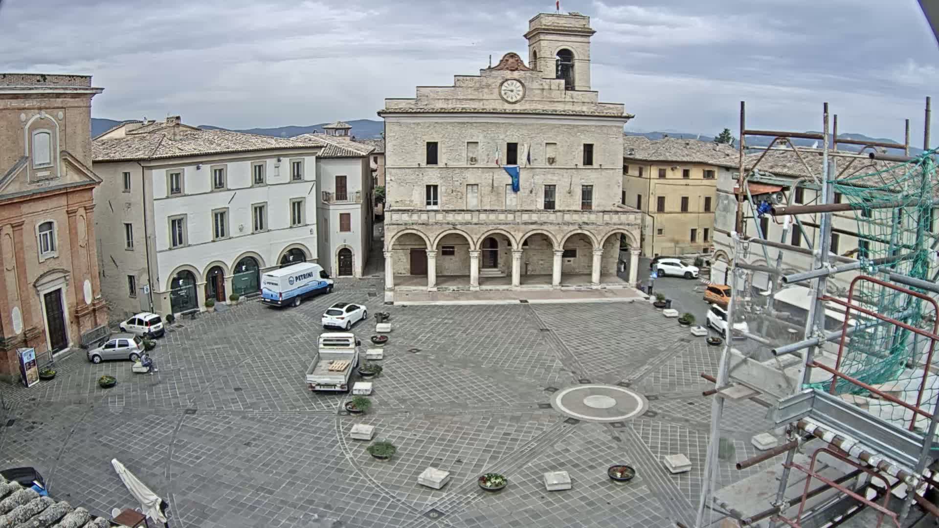 A town square with several buildings, including a large stone structure with arched entryways, is visible under an overcast sky, and a few cars are parked in the square.