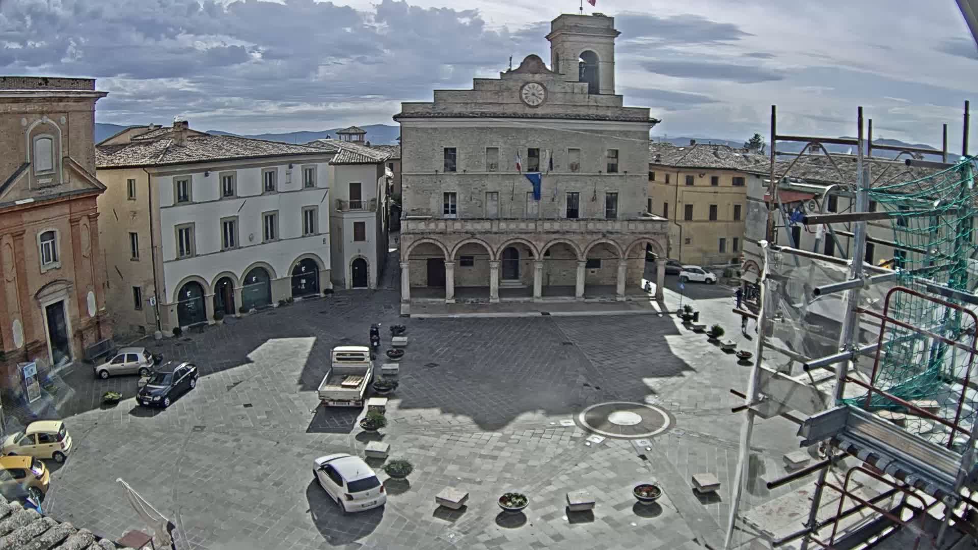 A town square, under partly cloudy skies, features a large stone building with arched entryways, surrounded by other buildings and a few cars.