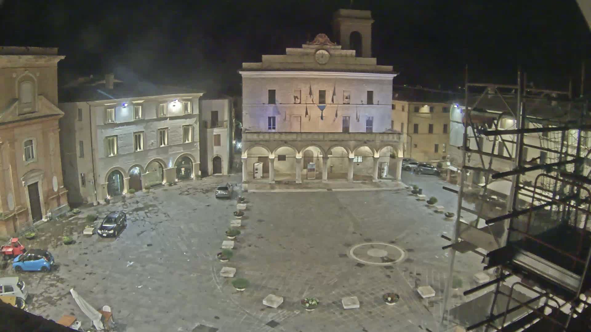 A nighttime view of a town square with several buildings surrounding it, including a large building with arched entryways, under slightly overcast conditions.