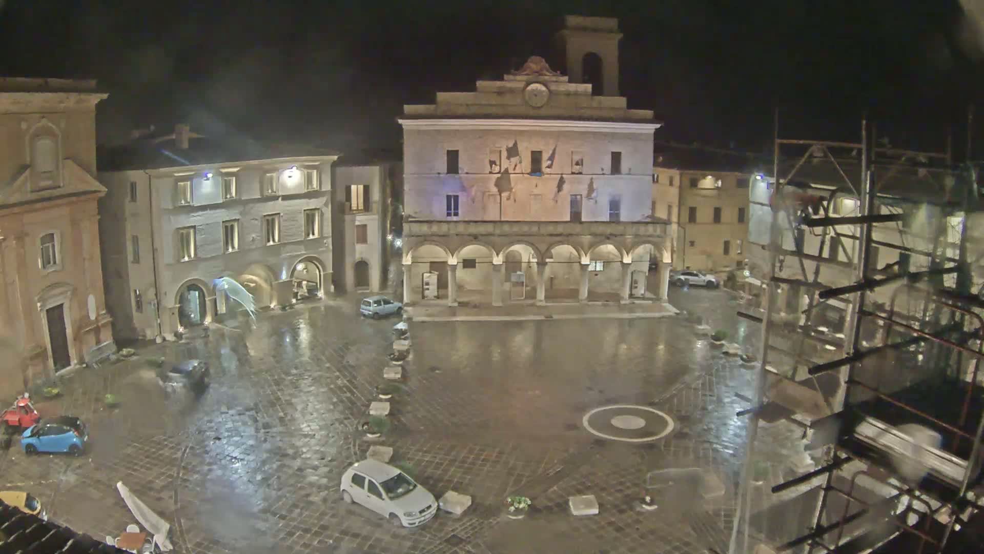 A rain-soaked town square at night, featuring several buildings, a few parked cars, and scaffolding in the lower right corner.
