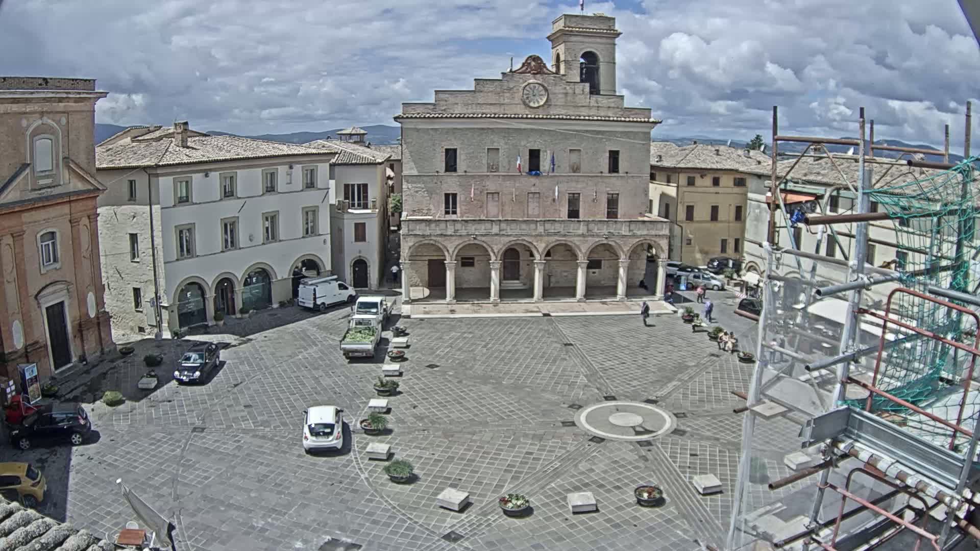 A town square, under partly cloudy skies, is centered around a large stone building with arched entryways, surrounded by other buildings and a few parked cars and pedestrians.