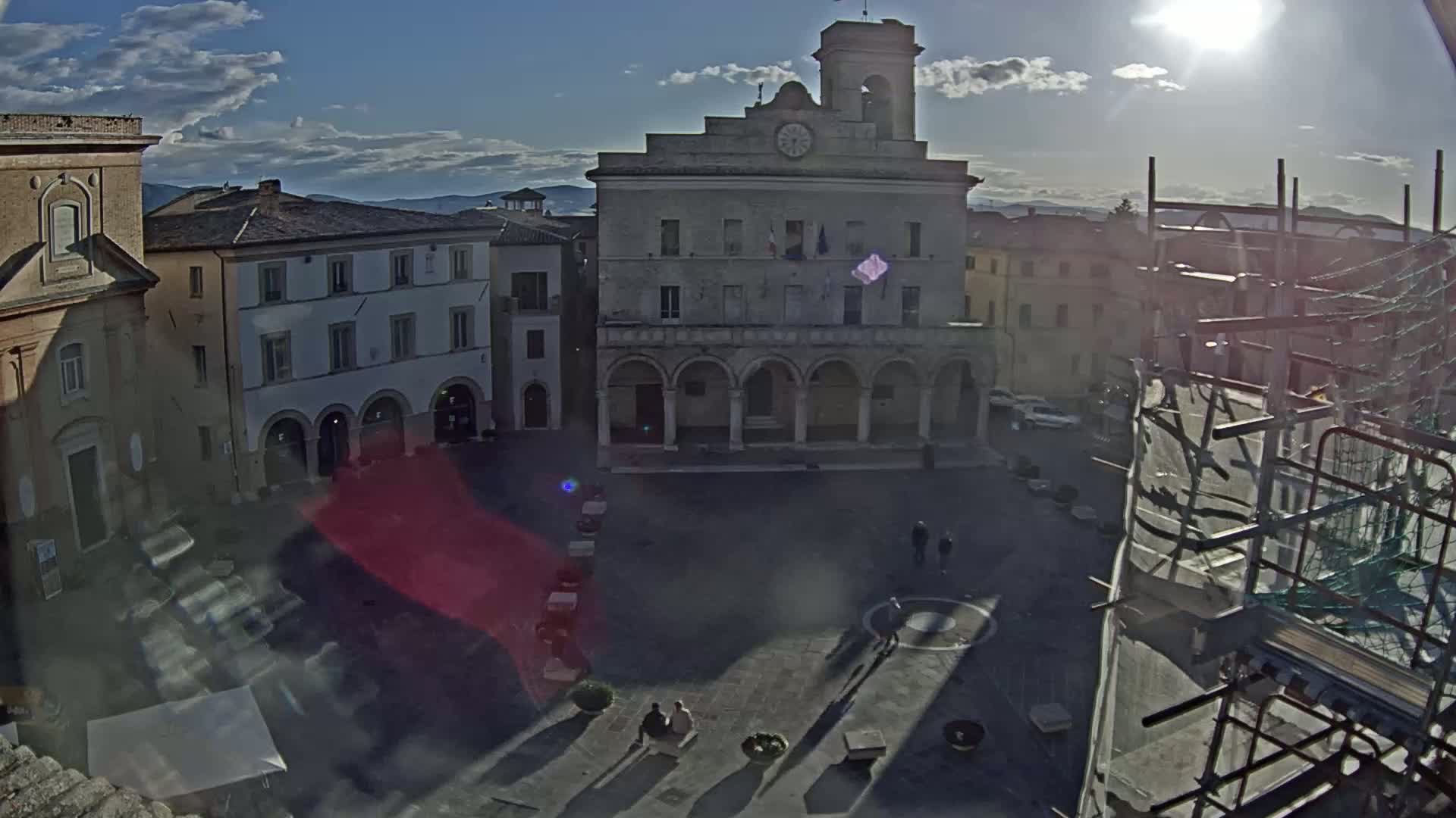 A sunny day reveals a large town square with several historic buildings, including a clock tower, and scaffolding obscuring part of the view.