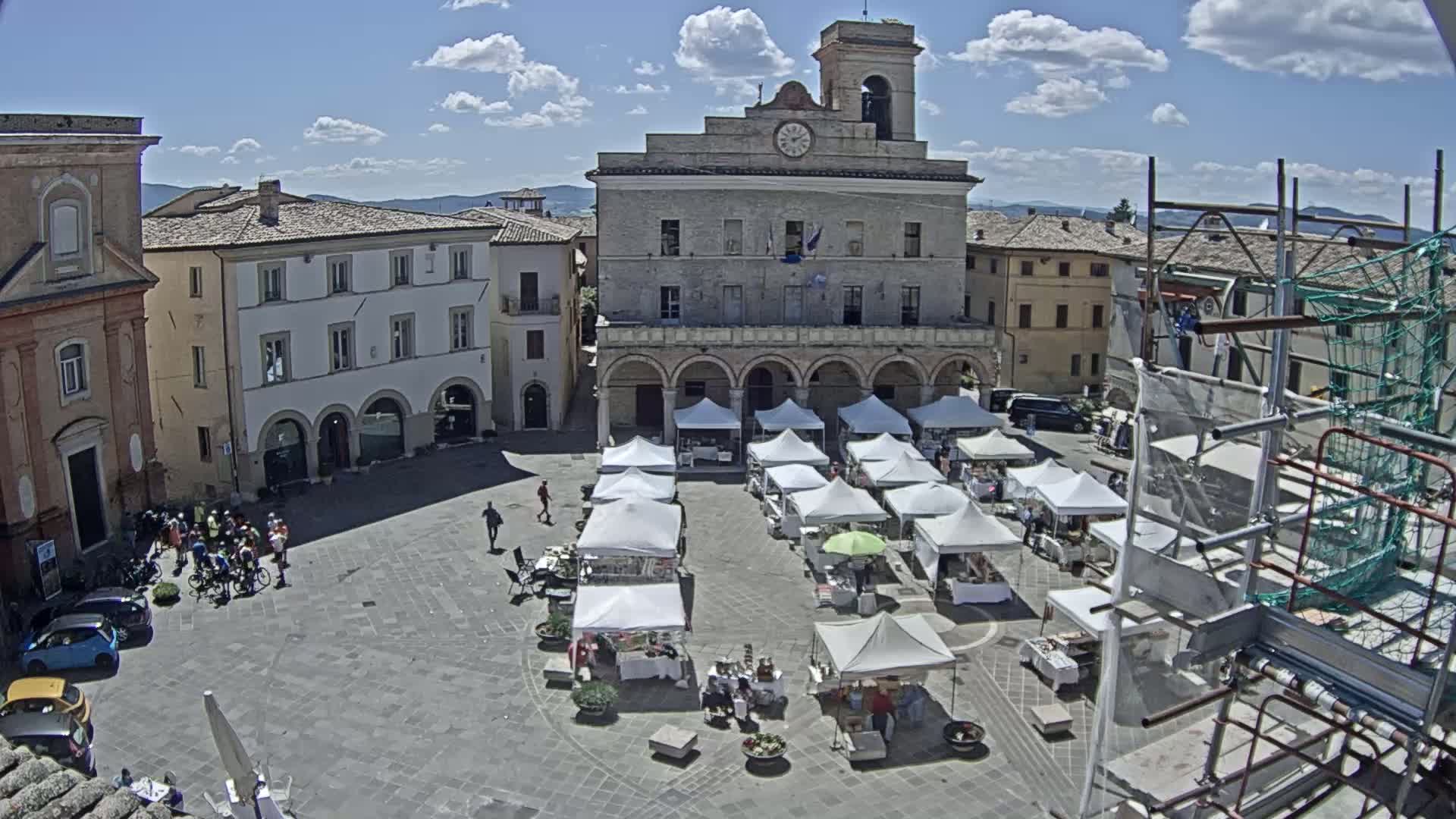 A sunny town square with numerous white market stalls set up in front of a large building with arched walkways, surrounded by other buildings.