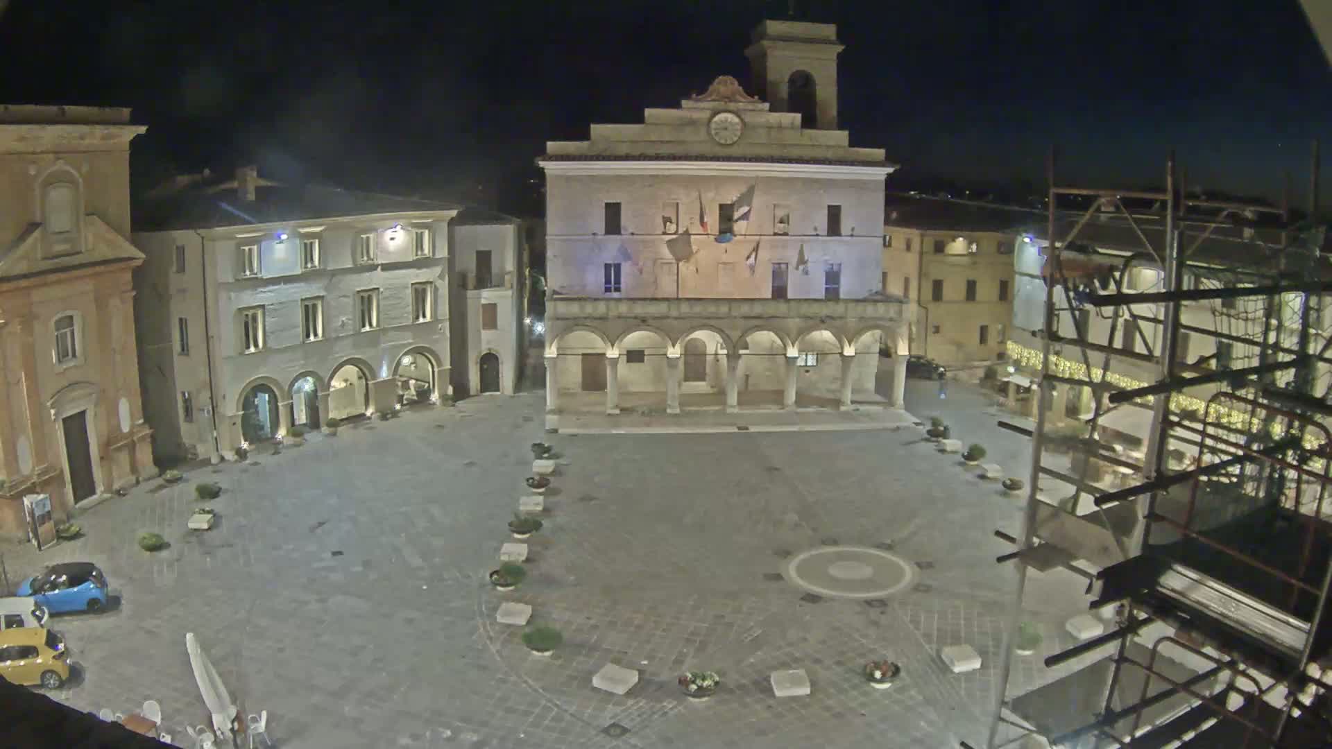 A nighttime view of a town square with several buildings, including a large building with arched entryways, under a clear sky.