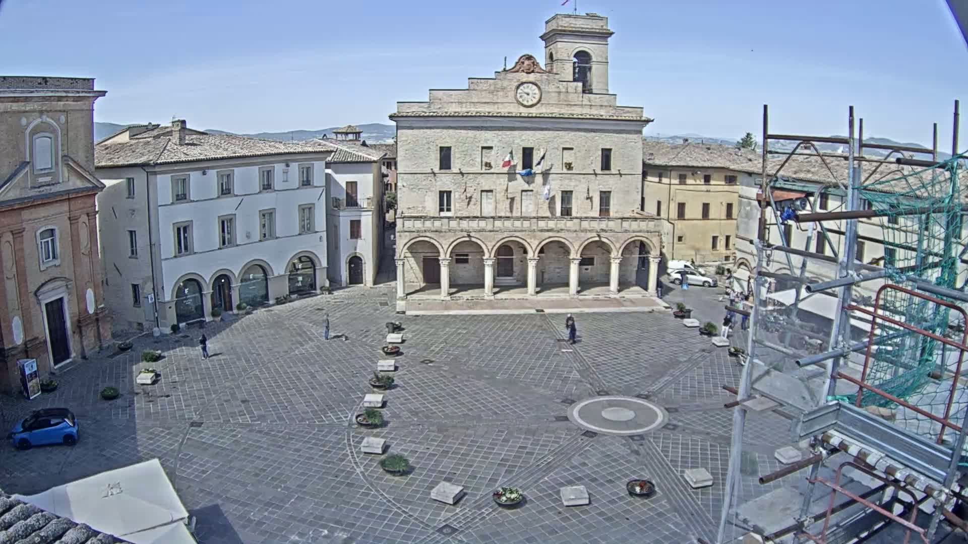 A large town square, under a clear blue sky, is centered on a large, arched building with a clock tower, surrounded by other buildings, and some scaffolding is visible in the lower right corner.