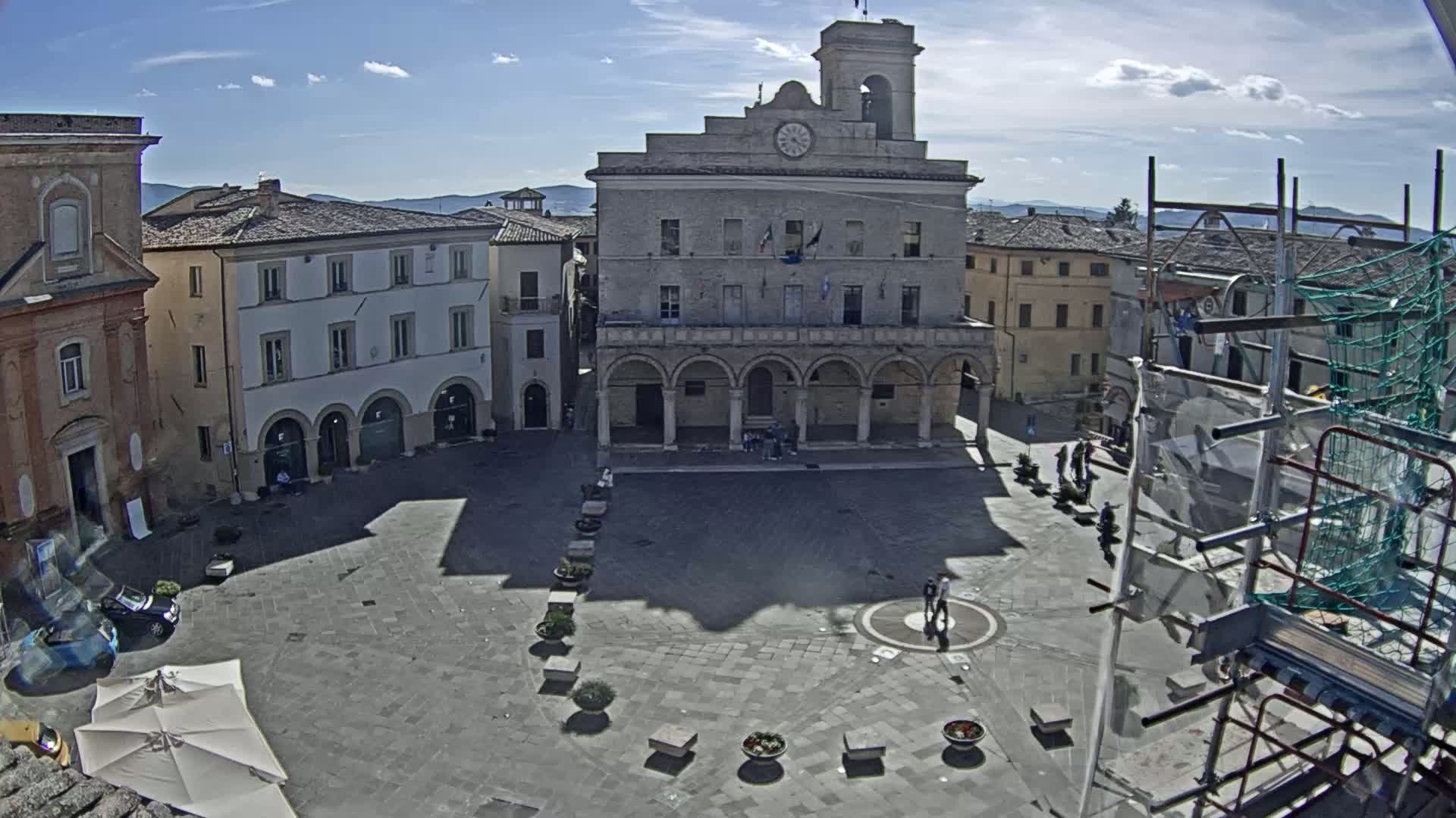 A town square with a large stone building, surrounded by other buildings, under a mostly sunny sky.