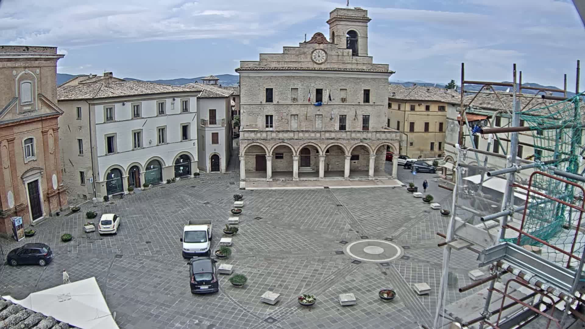 A town square with a large stone building featuring arched walkways, surrounded by other buildings, under a partly cloudy sky.