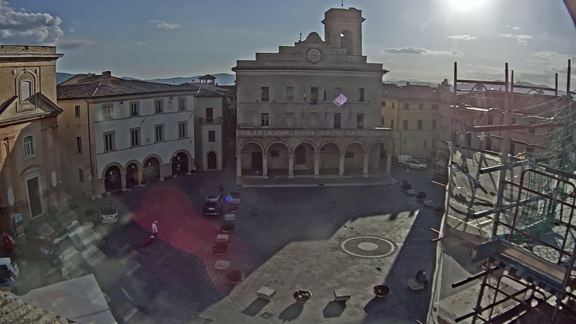 A sunny day reveals an expansive town square dominated by a large, classical-style building, with several other buildings and a few cars and people visible, and construction scaffolding on the right.
