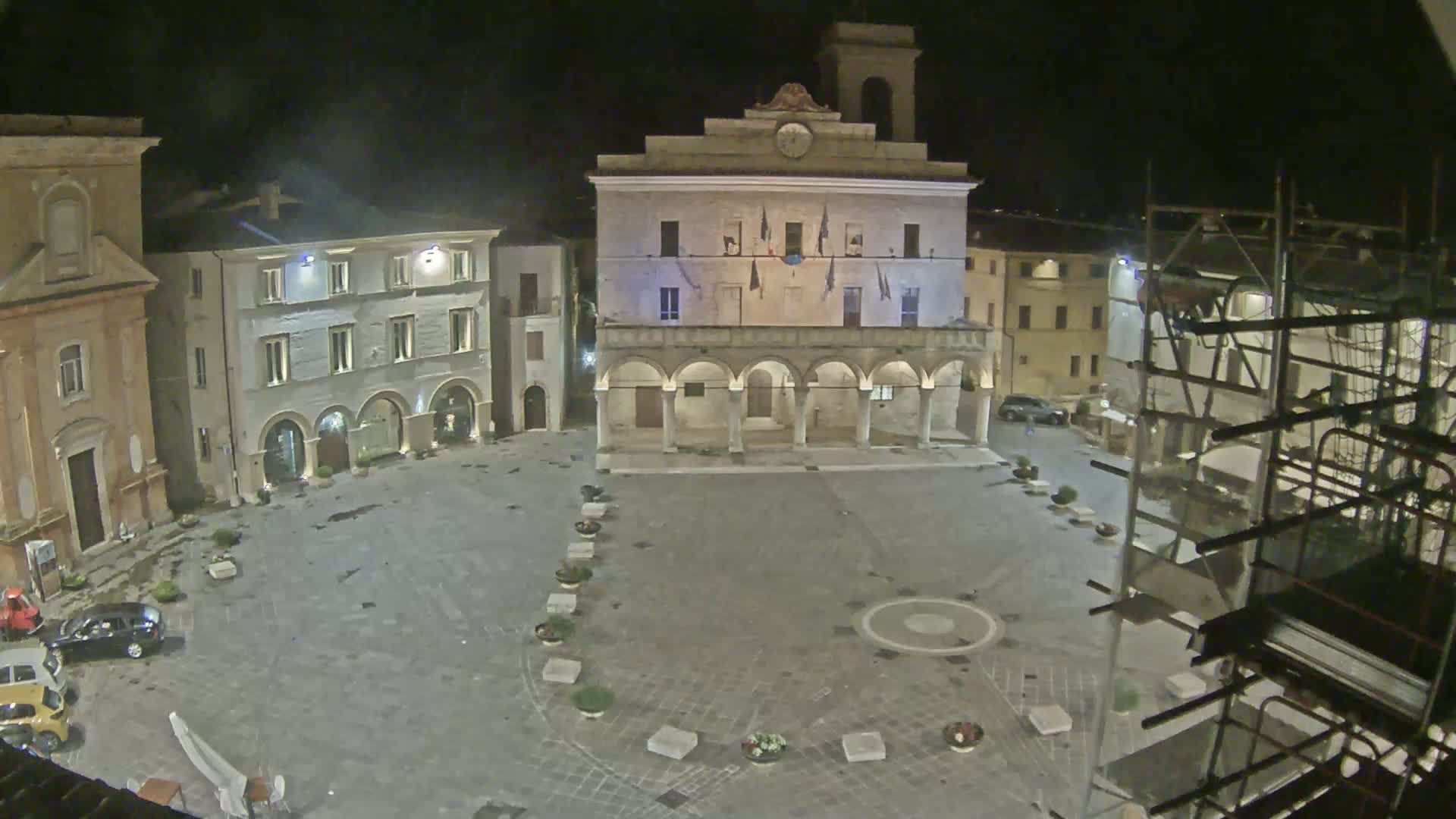 A nighttime view of a town square with a large, arched building in the center, surrounded by other buildings, under a clear, dark sky.