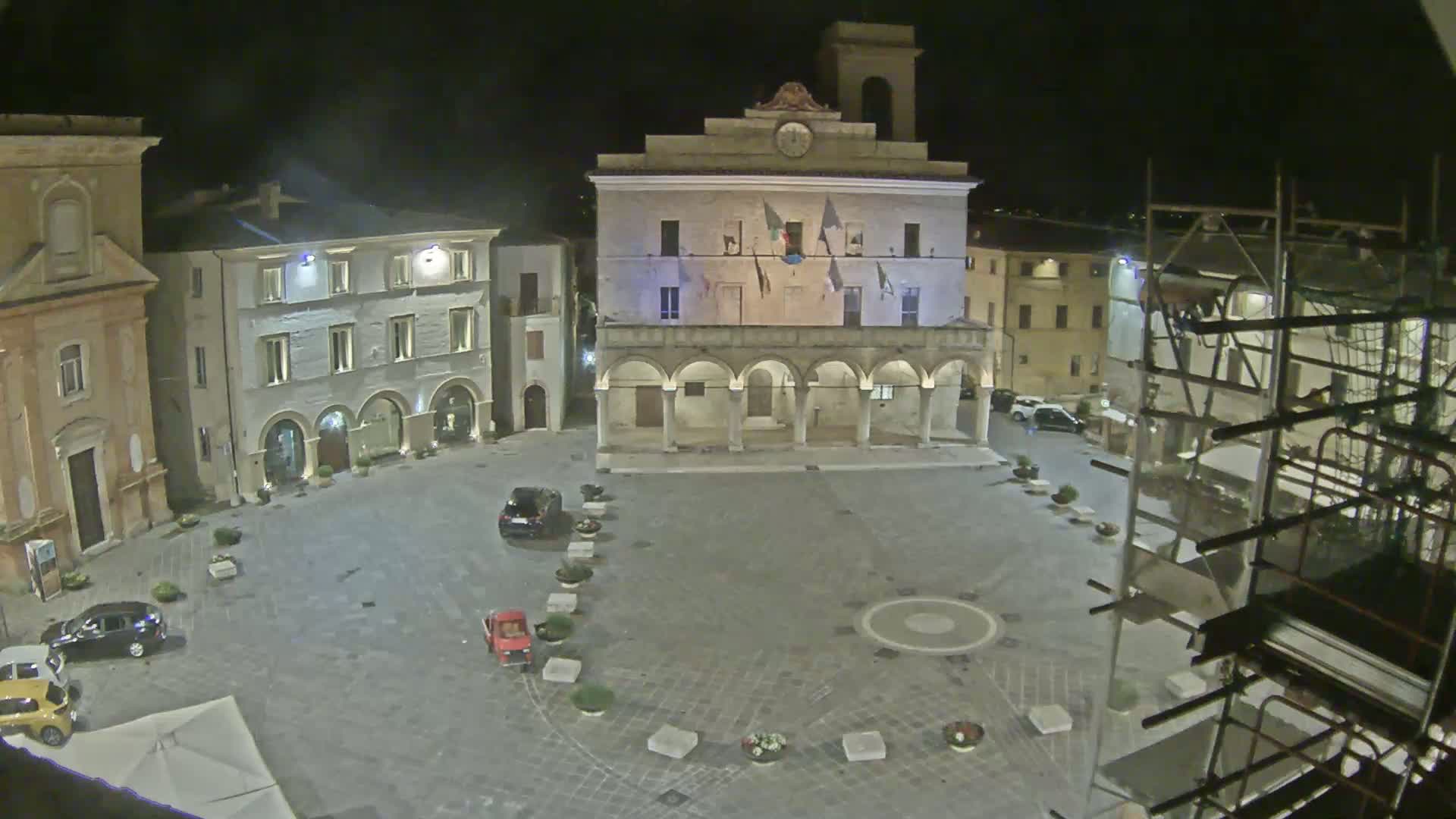 A nighttime view of a town square with a large building featuring arches, several cars parked in the square, and scaffolding visible in the lower right corner.