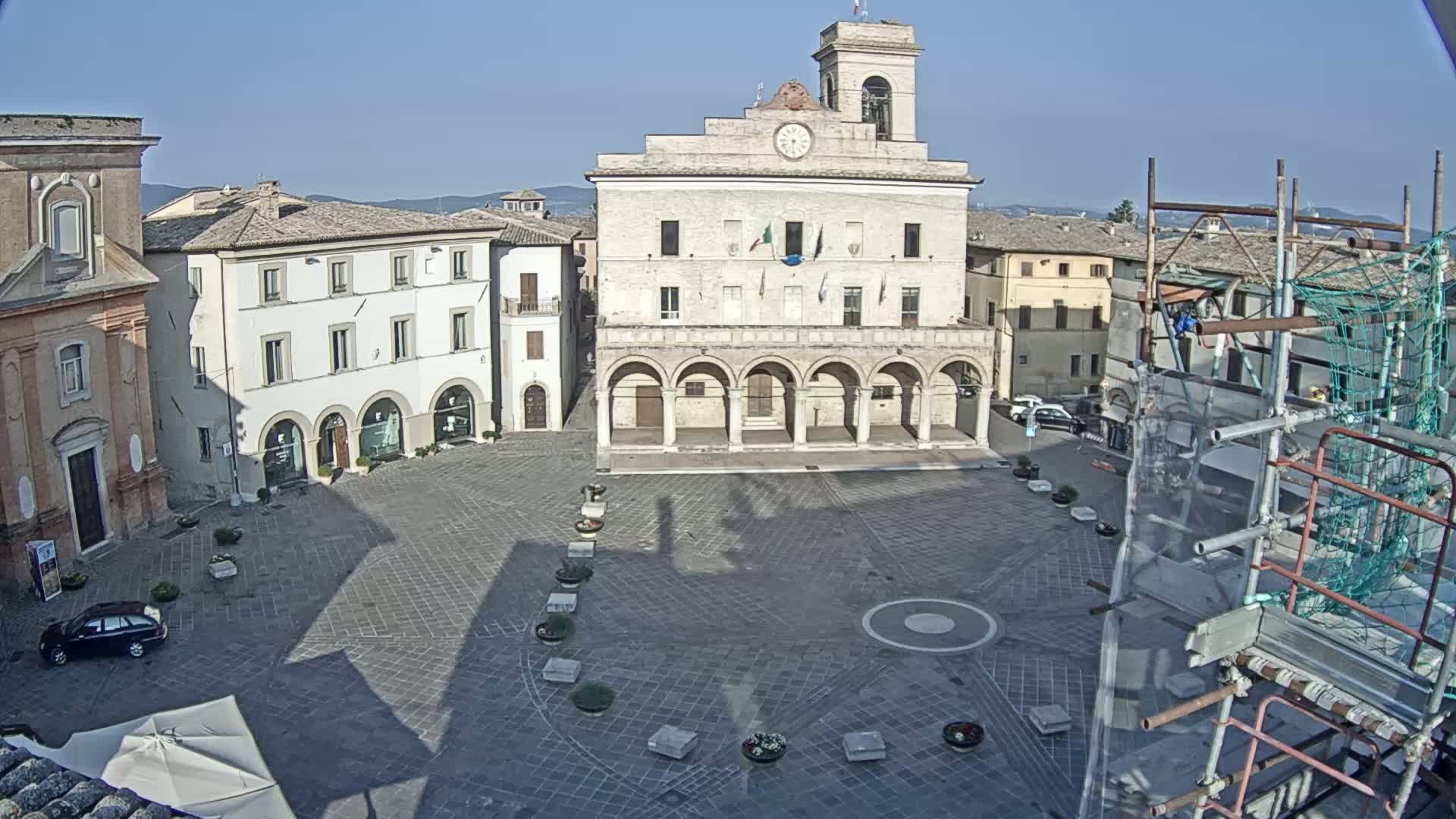 A large town square, paved in dark gray stone, is surrounded by buildings including a prominent stone structure with arched entryways and a clock tower, under a clear, sunny sky.