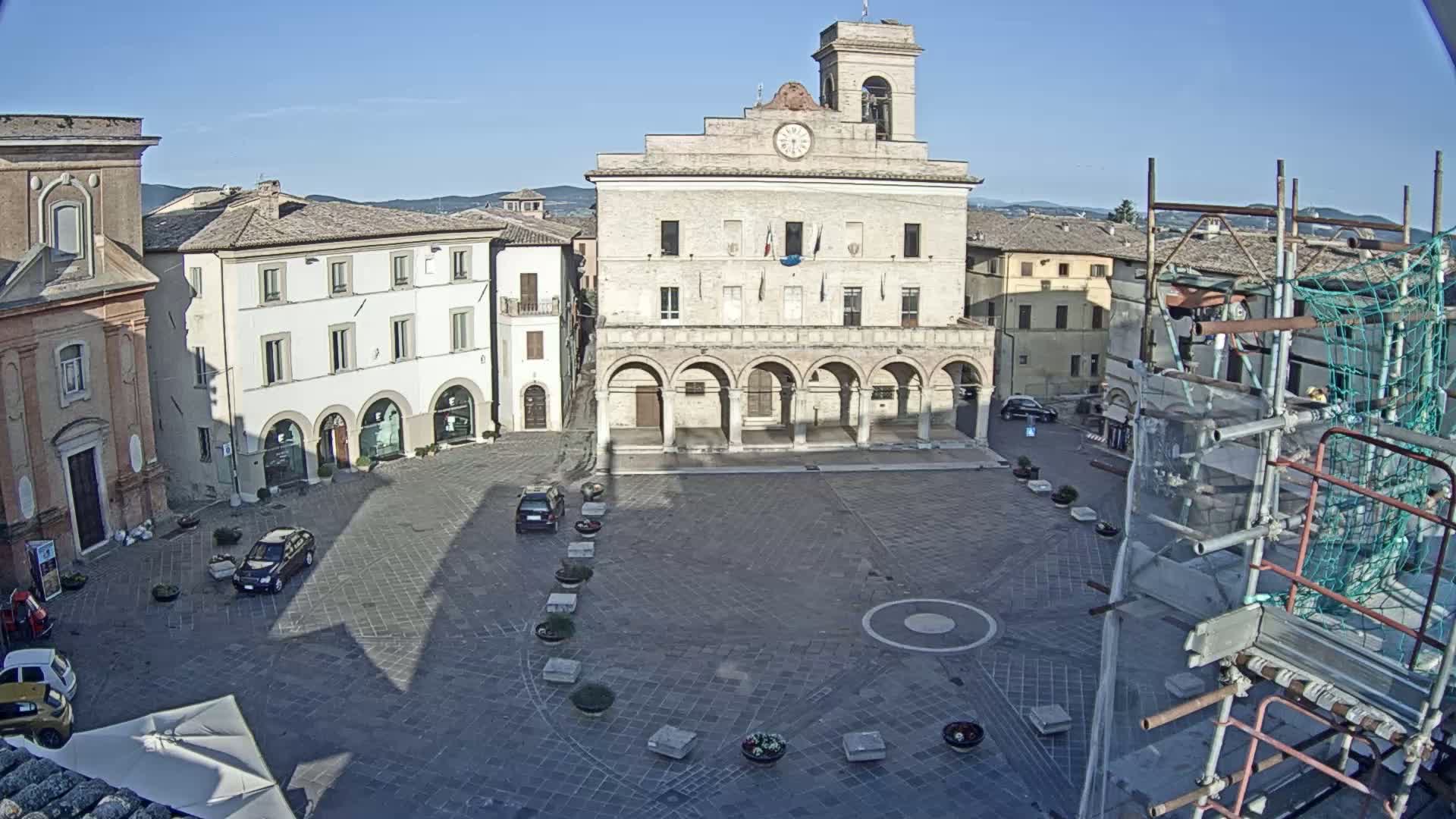 A large town square, under a clear blue sky, features a prominent stone building with arched entryways, surrounded by other buildings, some cars, and scaffolding.