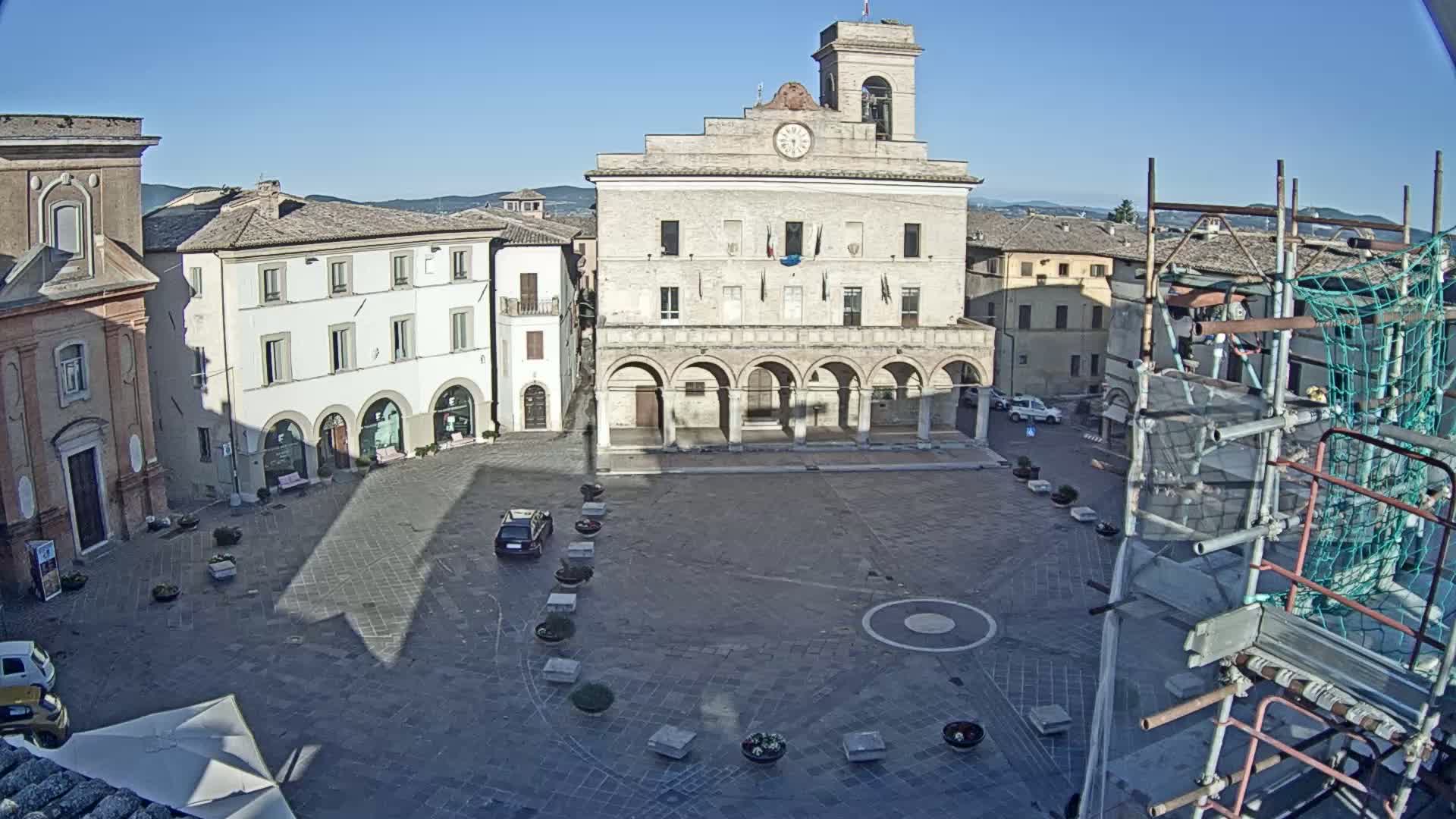 A large town square, under a clear blue sky, features a prominent stone building with arched walkways, surrounded by other buildings, and construction scaffolding is visible to the right.