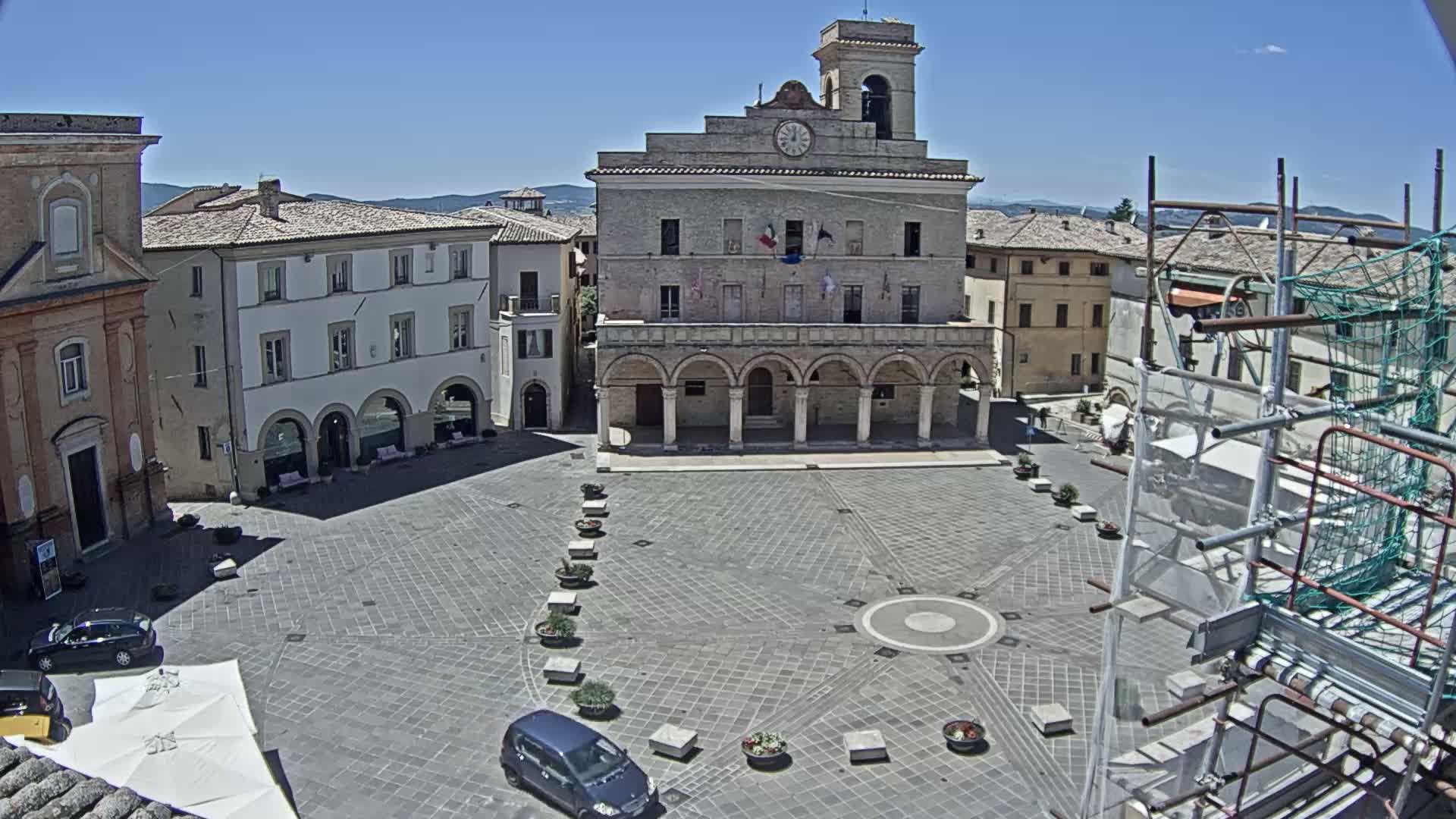 A town square with a large, pale stone building at its center, surrounded by other buildings, under a clear blue sky.