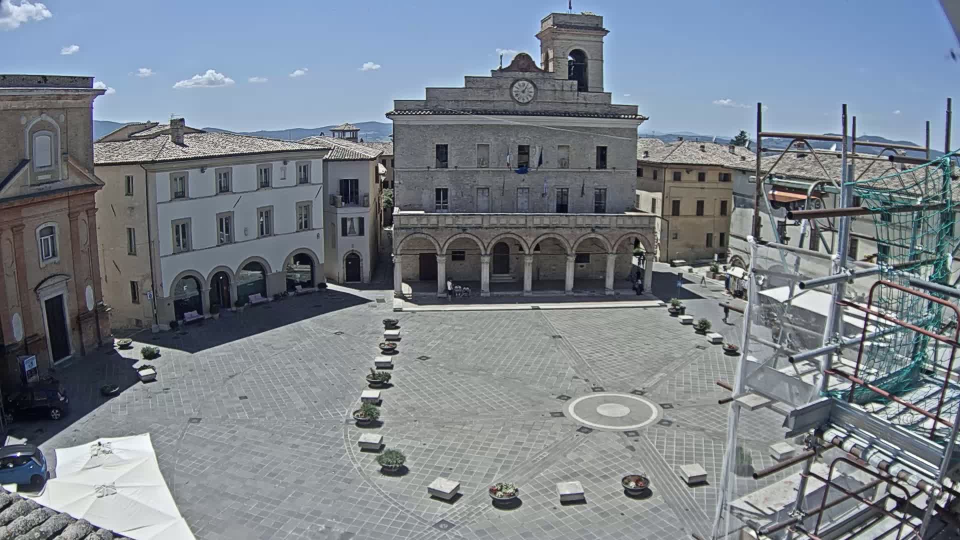 A town square with a large stone building and surrounding buildings, under a sunny sky with a few clouds.