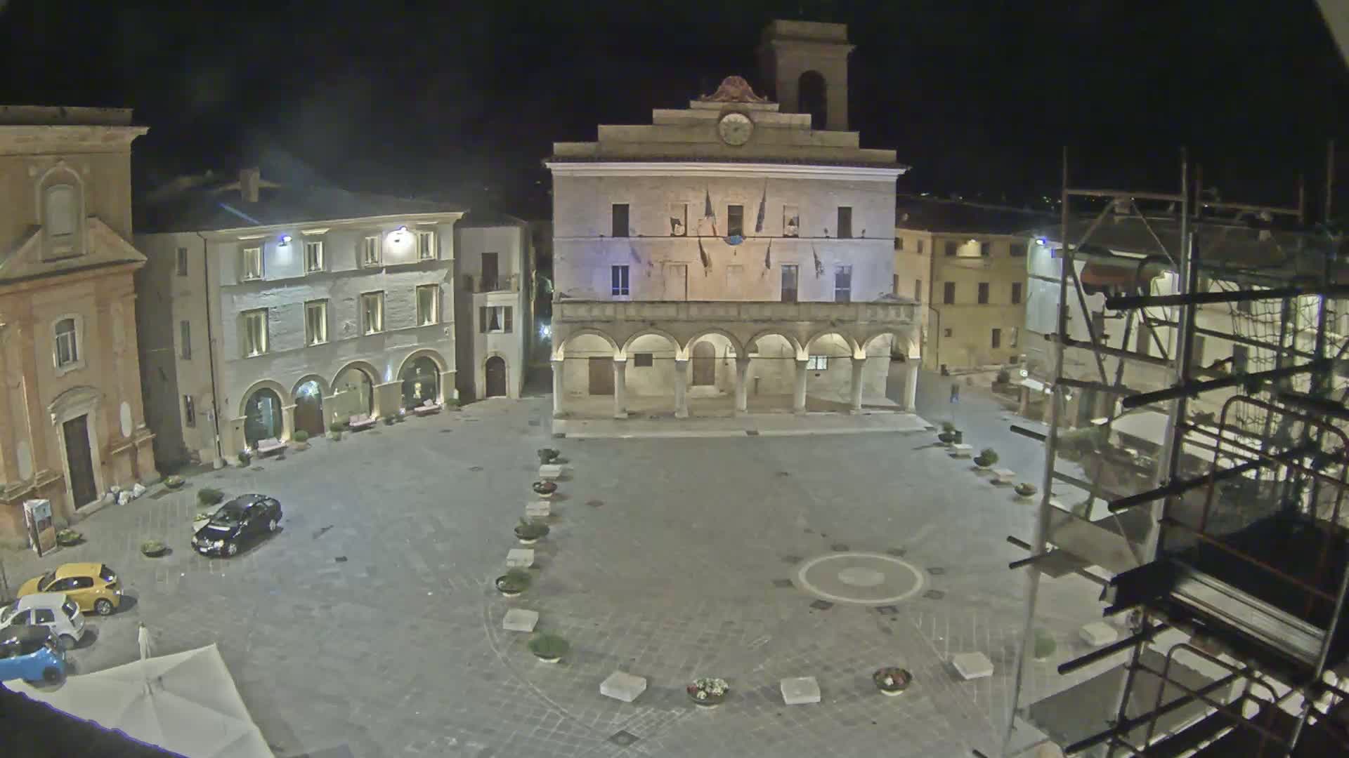 A nighttime view of a town square, illuminated, with several buildings surrounding it, including a large building with arches, and scaffolding on one side.