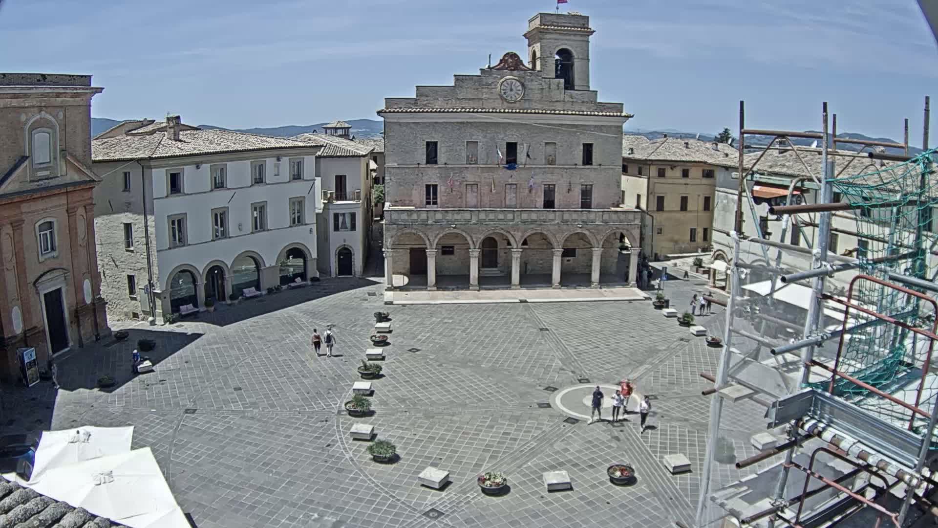 A large, stone town square with a prominent building featuring arched colonnades is visible under a bright, sunny sky.