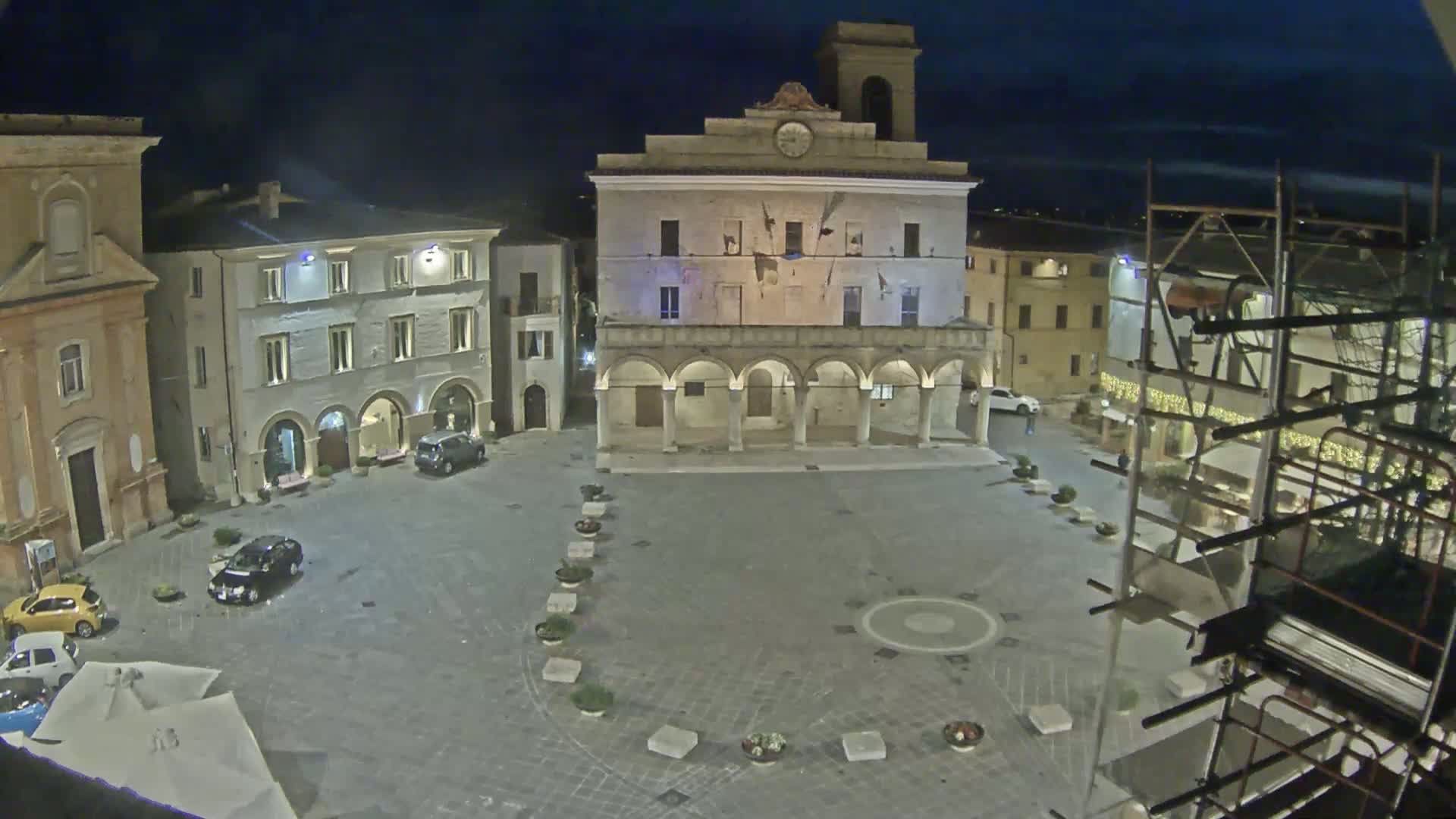 A nighttime view of a town square, centered around a large building with arched entryways, surrounded by other buildings under a dark, clear sky.