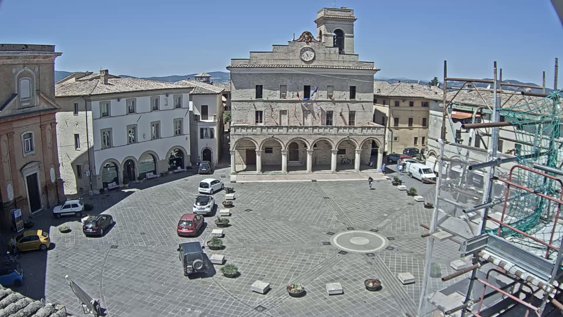 A town square with a large building, several parked cars, and scaffolding, on a sunny day.