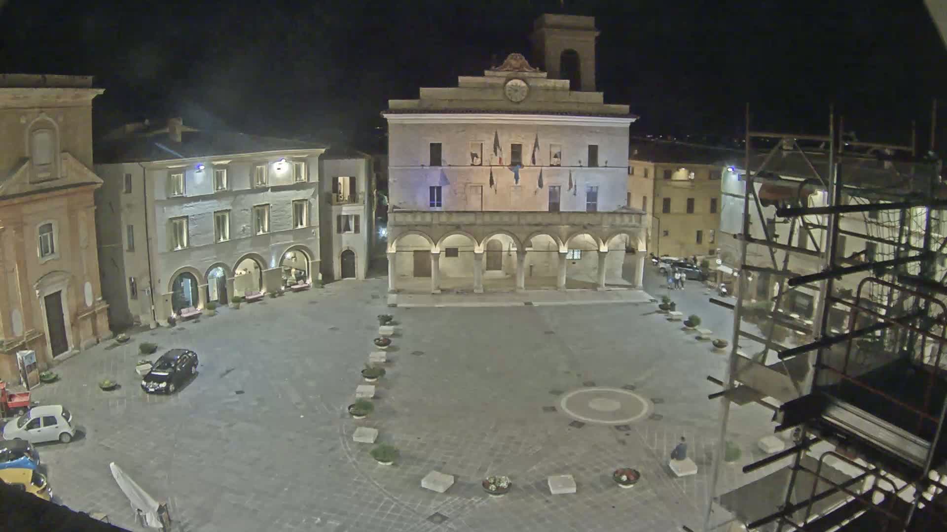 A nighttime, high-angle view of a town square with a large building featuring arched walkways, surrounded by other buildings, a few cars, and scaffolding.