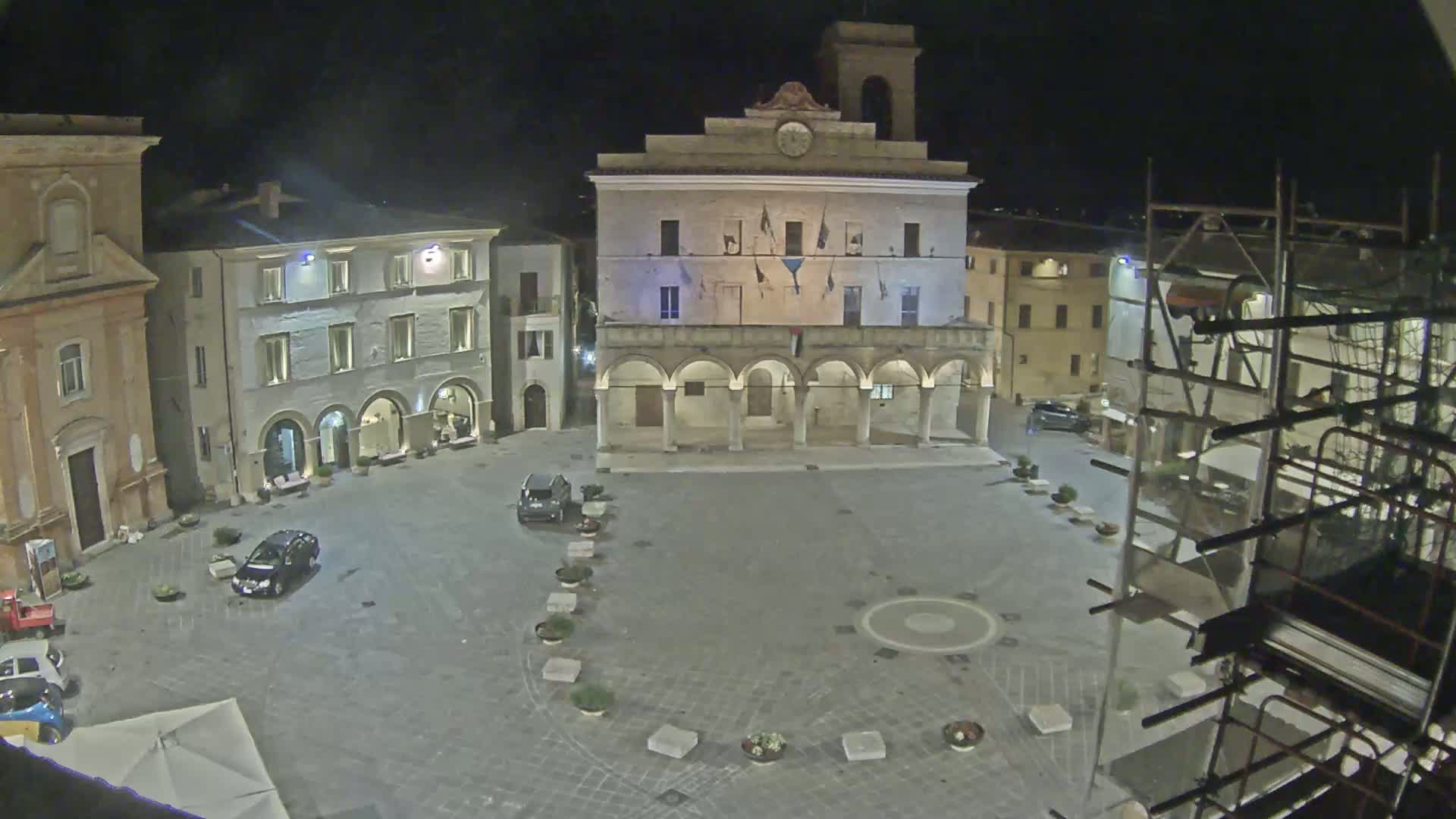 A nighttime view of a town square, featuring a large building with arched entryways, several other buildings, and a few cars parked in the square.
