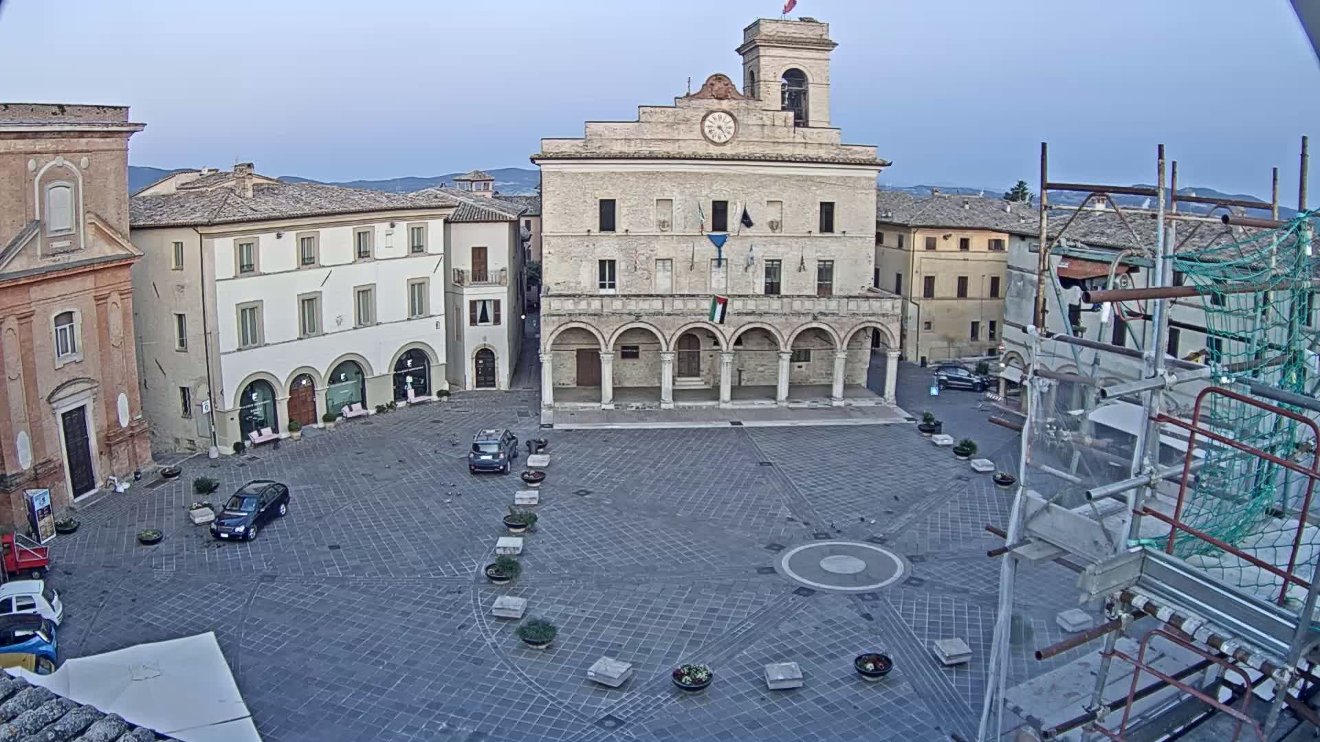 A town square with a large stone building, several smaller buildings, and a few cars under a clear, light-blue sky.