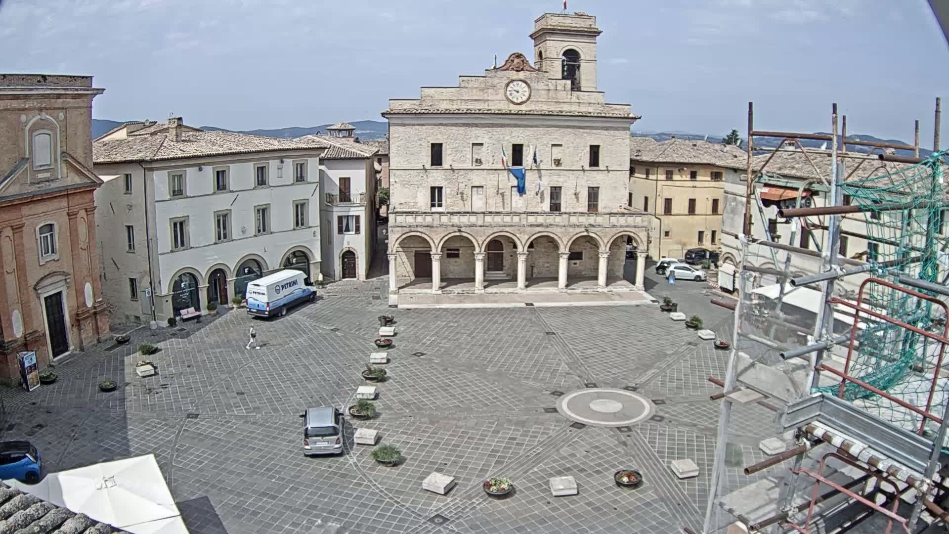 A large town square with a stone building featuring arched entrances, several other buildings, a few cars, and scaffolding, all under a clear, sunny sky.