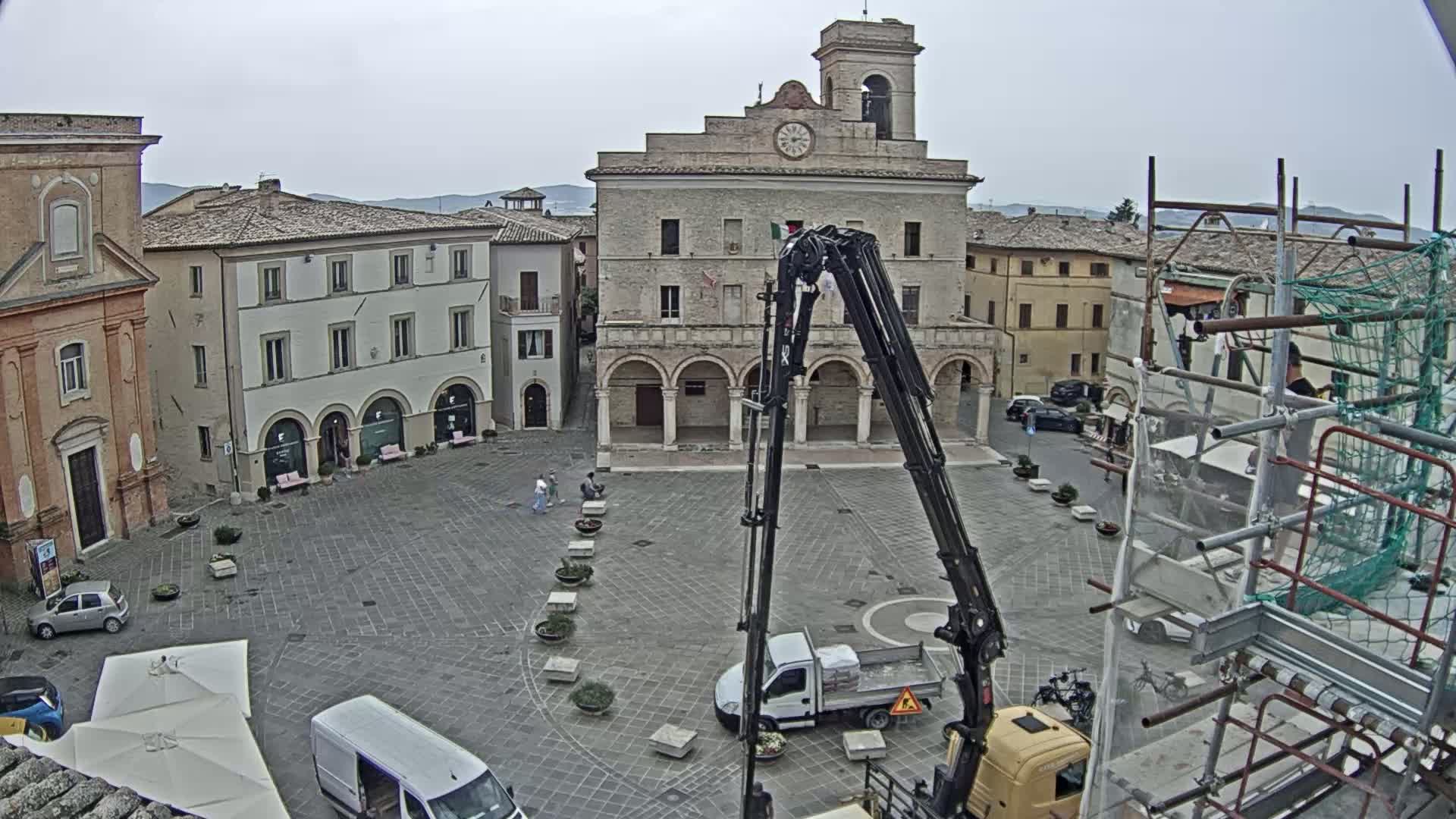 An overcast day reveals a town square with several buildings, including a large stone structure with arched entryways, a crane and truck, and scaffolding surrounding a building under construction.