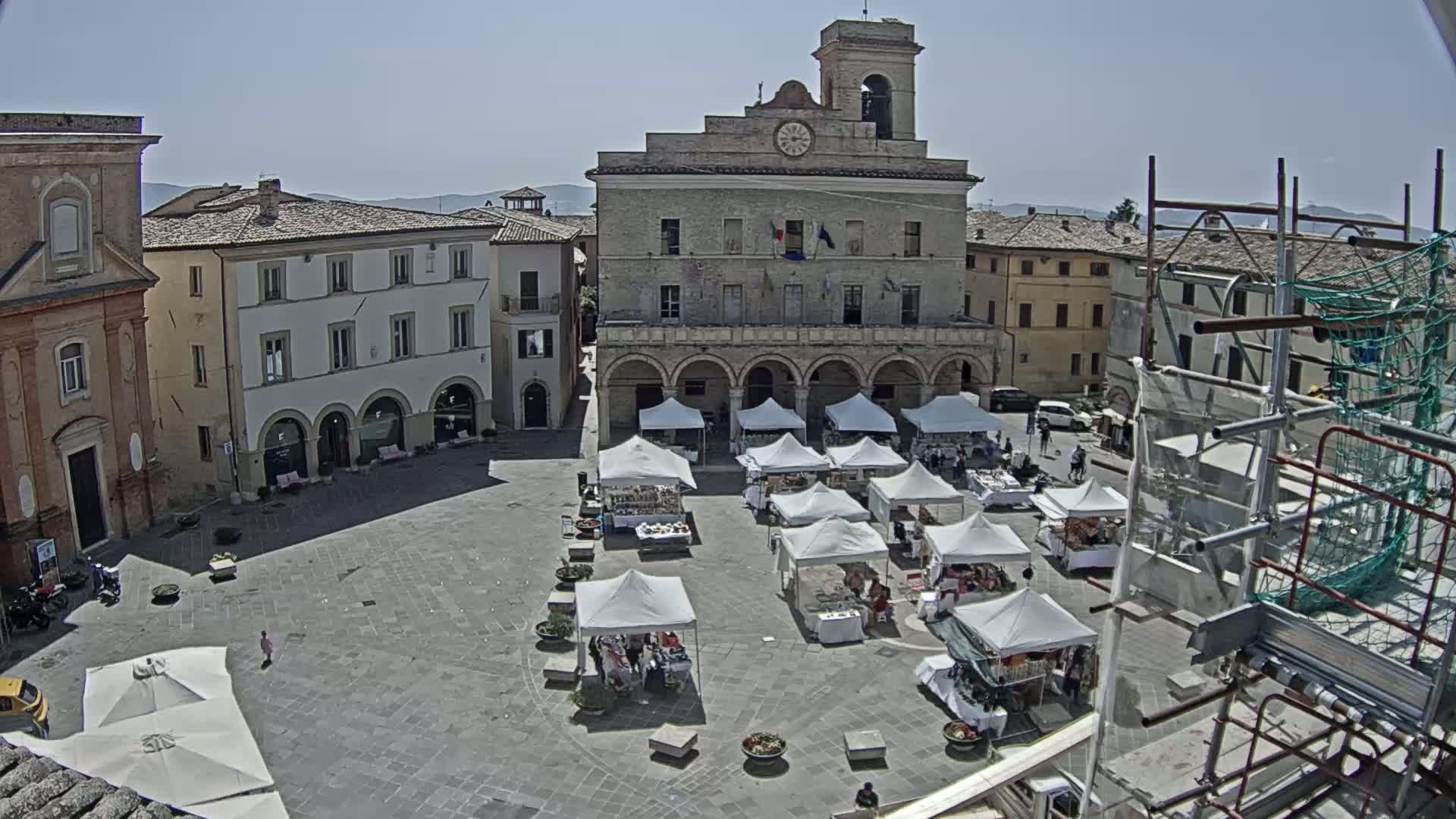 A sunny day reveals a town square with numerous market stalls under white canopies, surrounded by historic buildings, including a large, arcaded town hall.